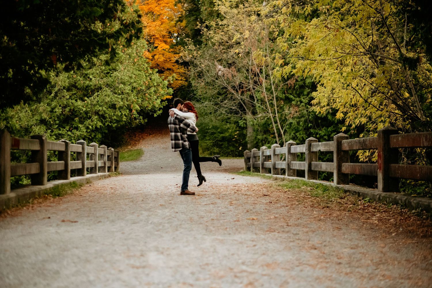A figure walks along a wooden bridge pathway surrounded by vibrant autumn foliage in a peaceful park setting.