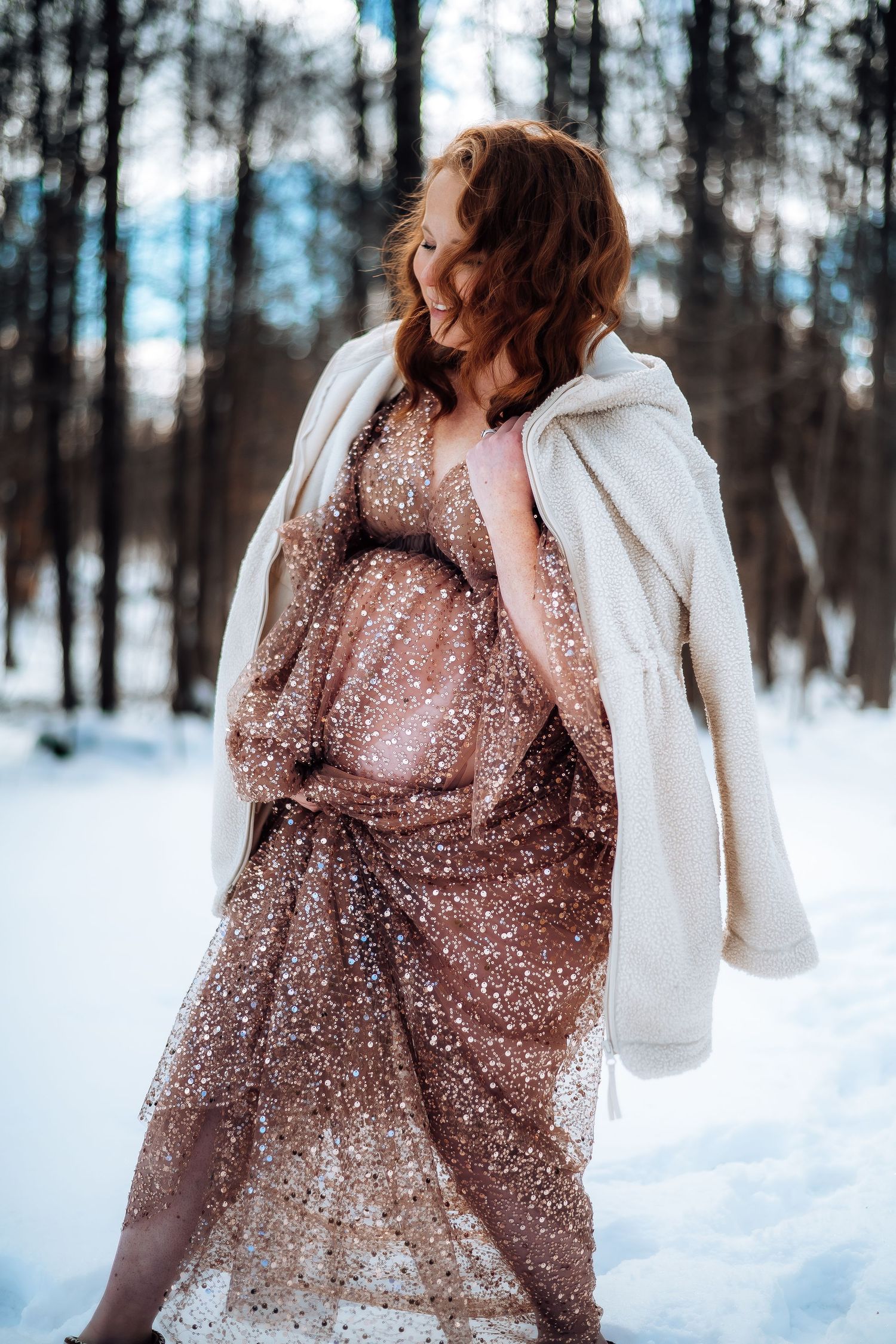 Winter maternity photo featuring sparkly dress and white coat in snowy forest setting. Maternity photographer Ontario
