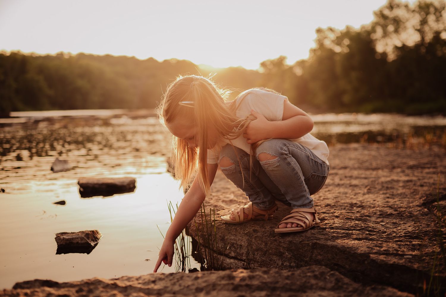 Silhouetted figures sit along a riverbank at sunset with warm golden light reflecting off the water's surface.