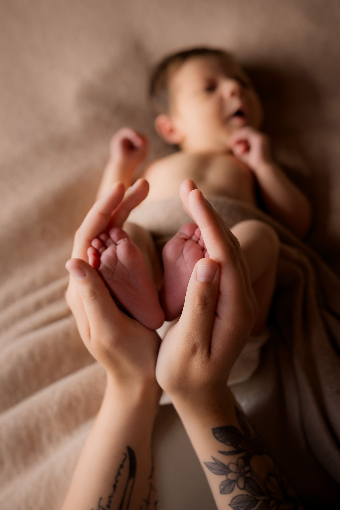 Tattooed hands gently cradling tiny newborn baby feet, with infant lying on soft beige blanket in background.