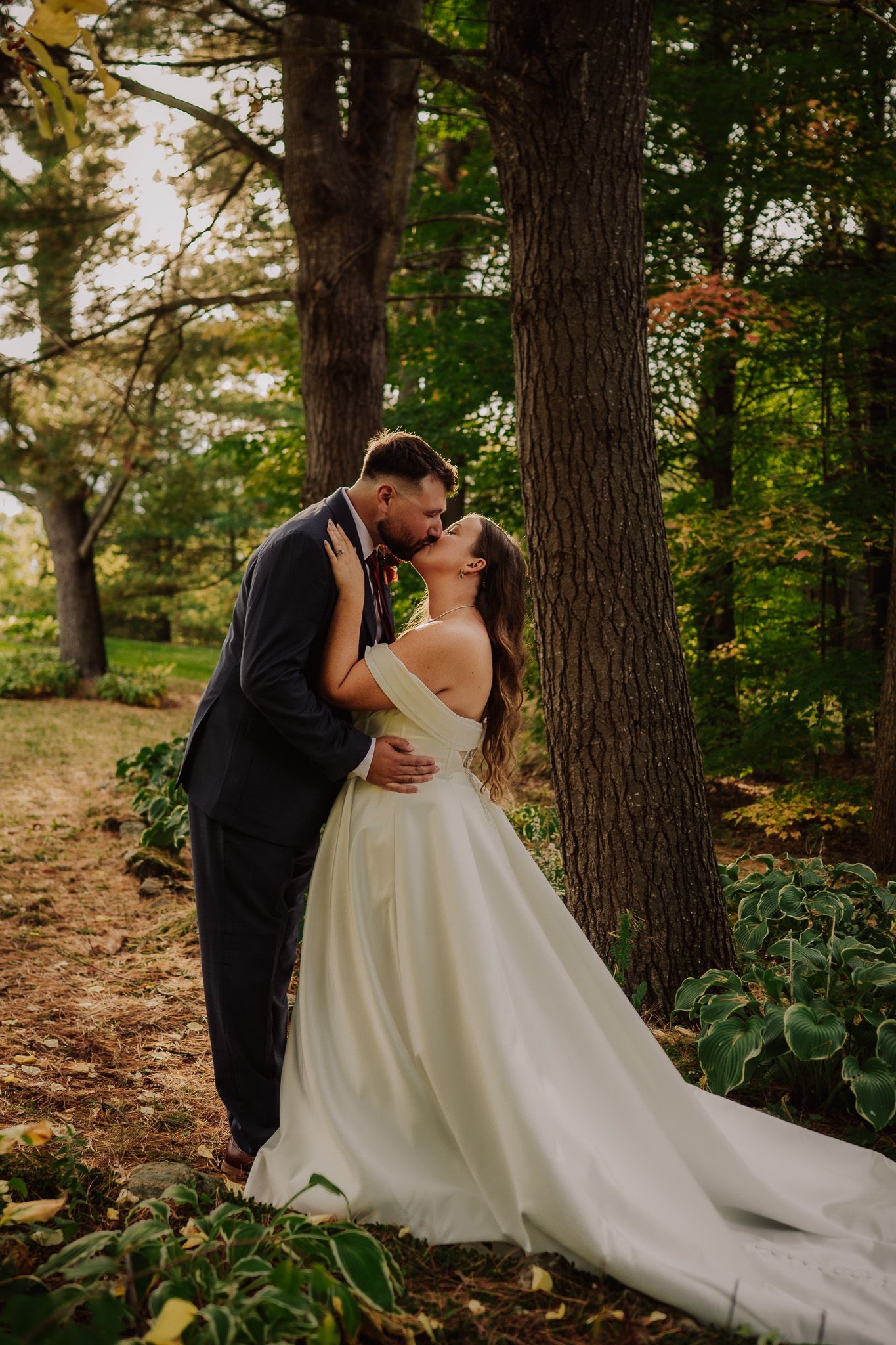A romantic wedding couple shares a passionate kiss in a wooded forest setting with a flowing white gown trailing behind them.