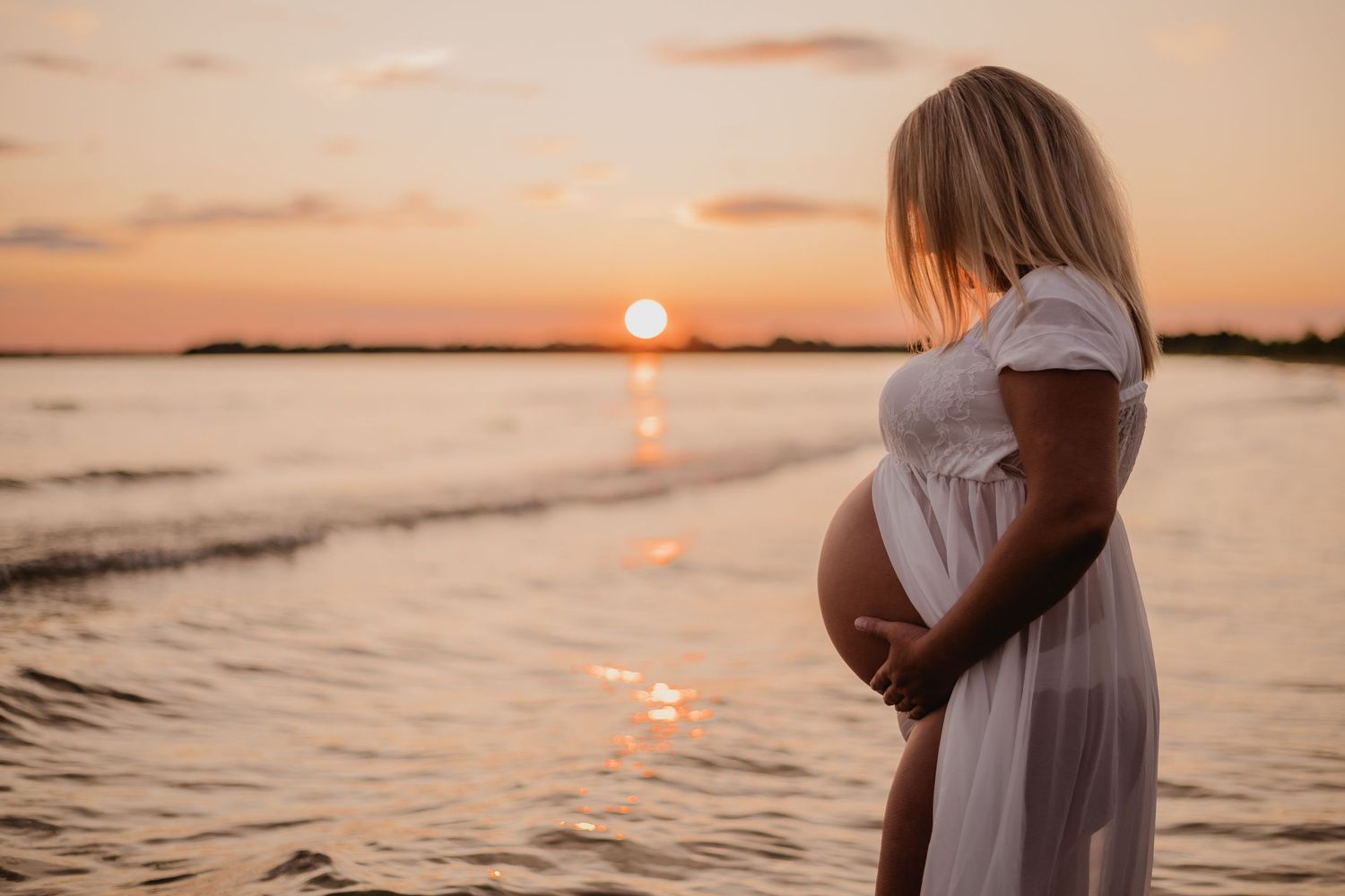 A flowing white dress silhouetted against a golden sunset over calm waters during a maternity photoshoot. Maternity photographer Ontario