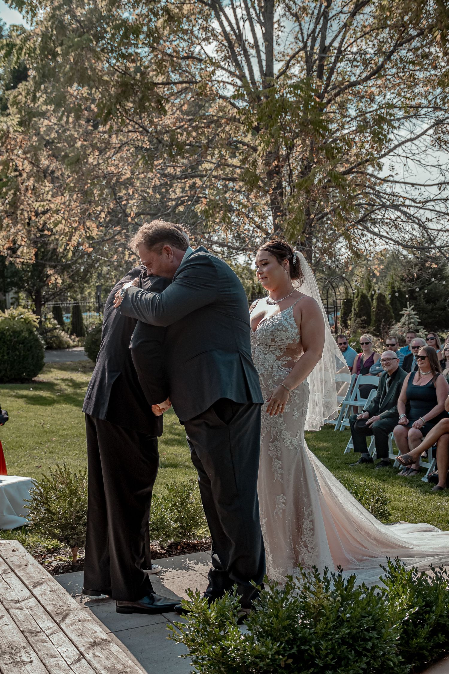 An emotional moment during an outdoor garden wedding ceremony with attendees seated on white chairs among landscaped grounds.