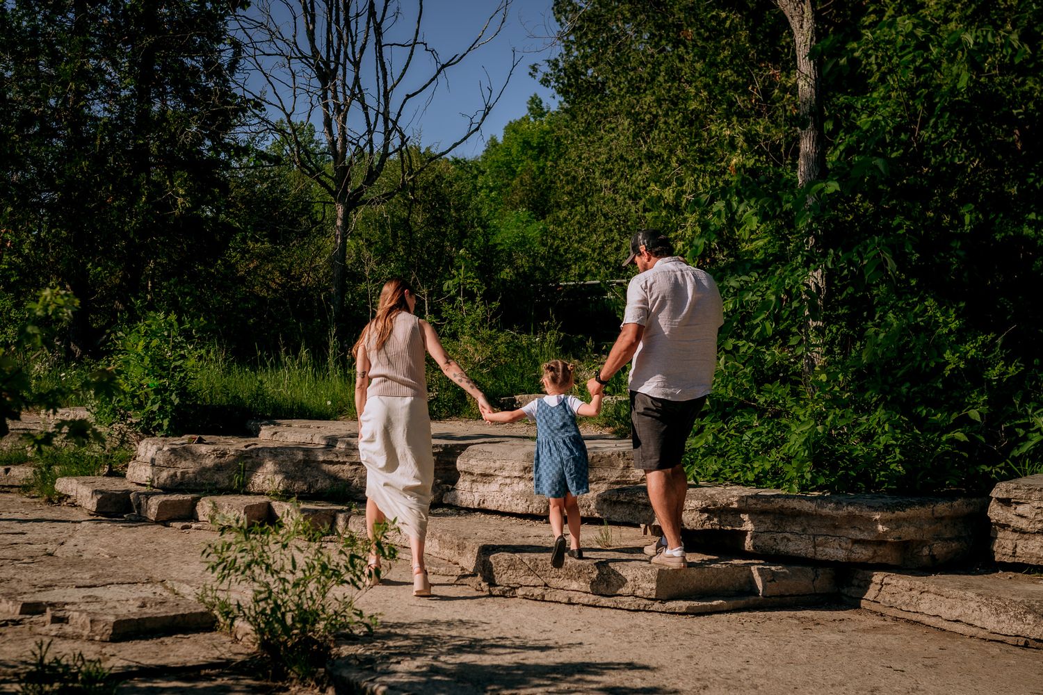 Family of three holding hands walking on stone steps surrounded by lush green trees in a sunny park.