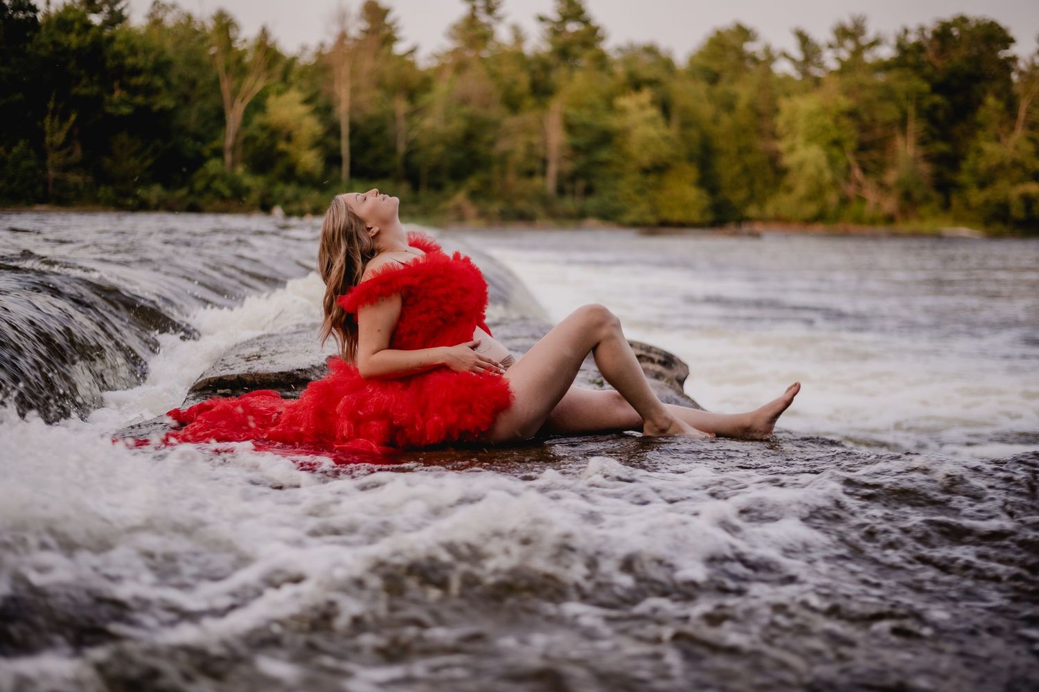 A red gown flows dramatically as water cascades over rocks during a waterfall photoshoot. Maternity photographer Ontario