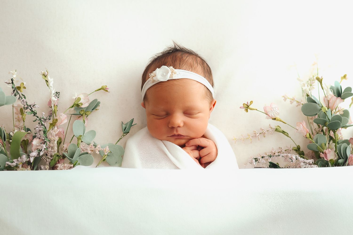 A sleeping newborn with a delicate headband poses among soft florals on mint green fabric.