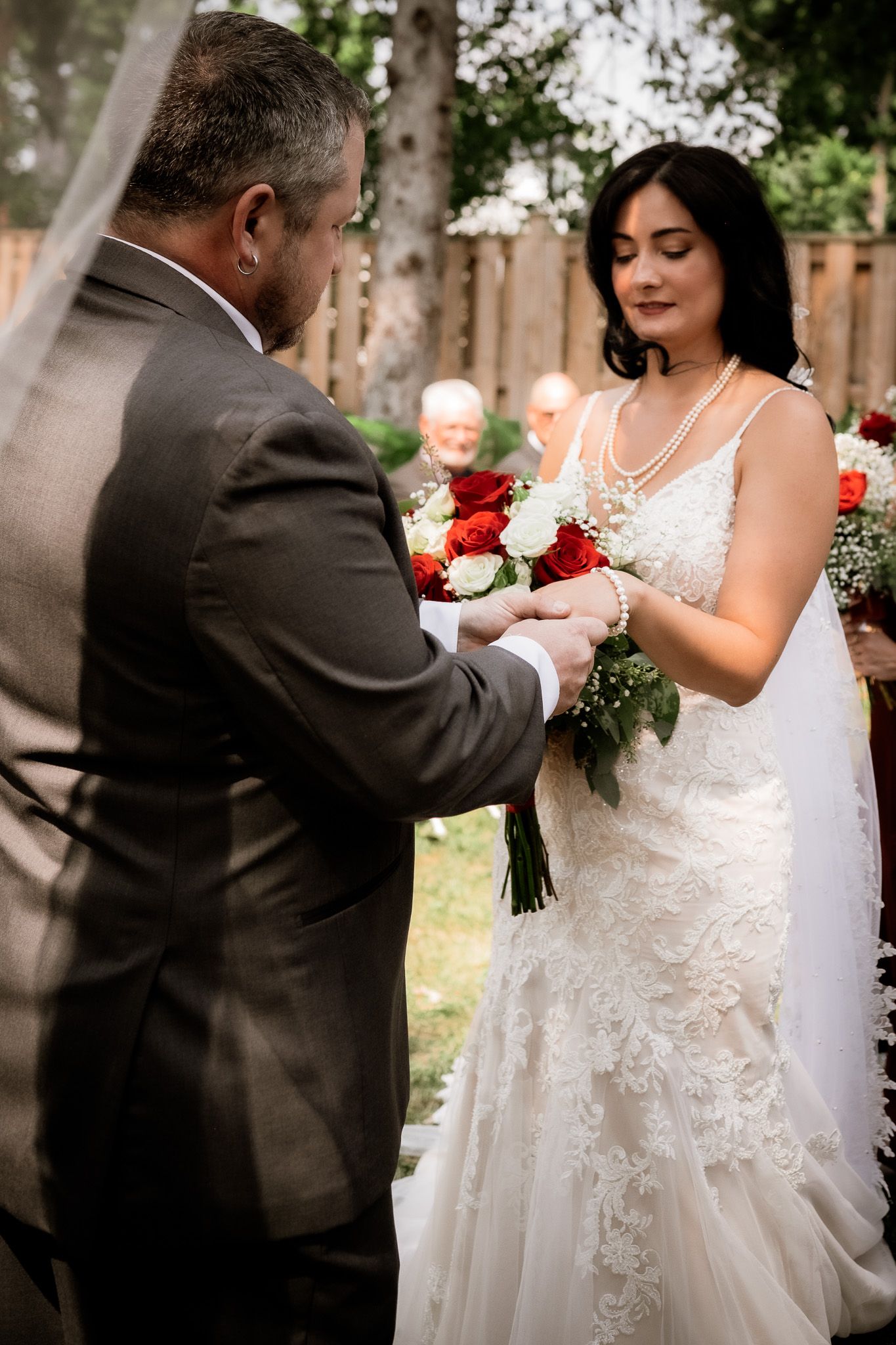 A couple in wedding attire exchange rings during an outdoor ceremony surrounded by greenery.