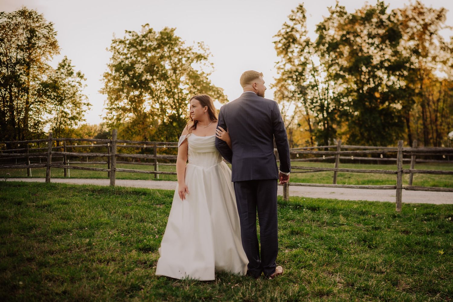 A couple in wedding attire stands together on a grassy lawn at sunset with wooden fencing and trees in the background.