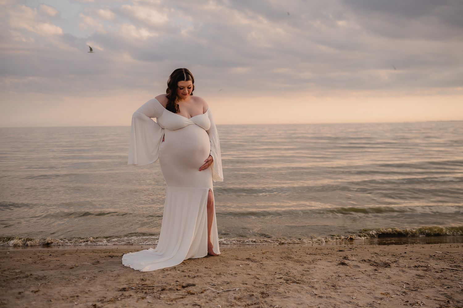 Expectant mother poses on beach during sunset in white flowing gown. Maternity photographer Ontario