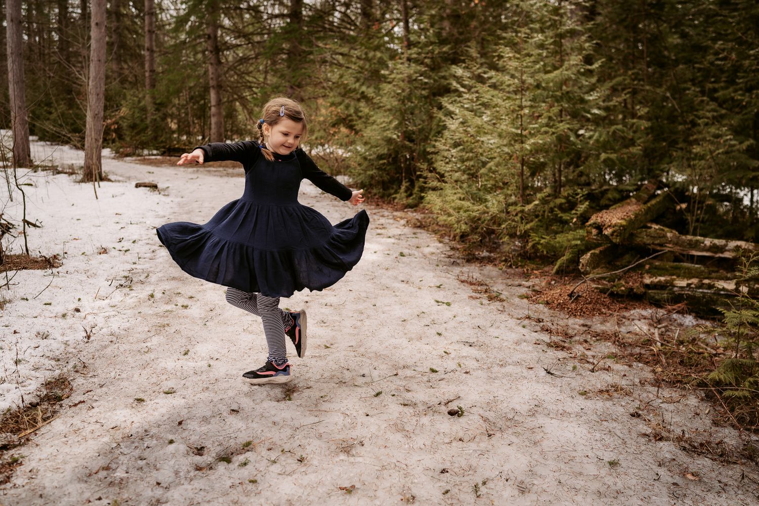 A young girl in a navy dress twirls joyfully on a snowy forest path surrounded by tall pine trees.