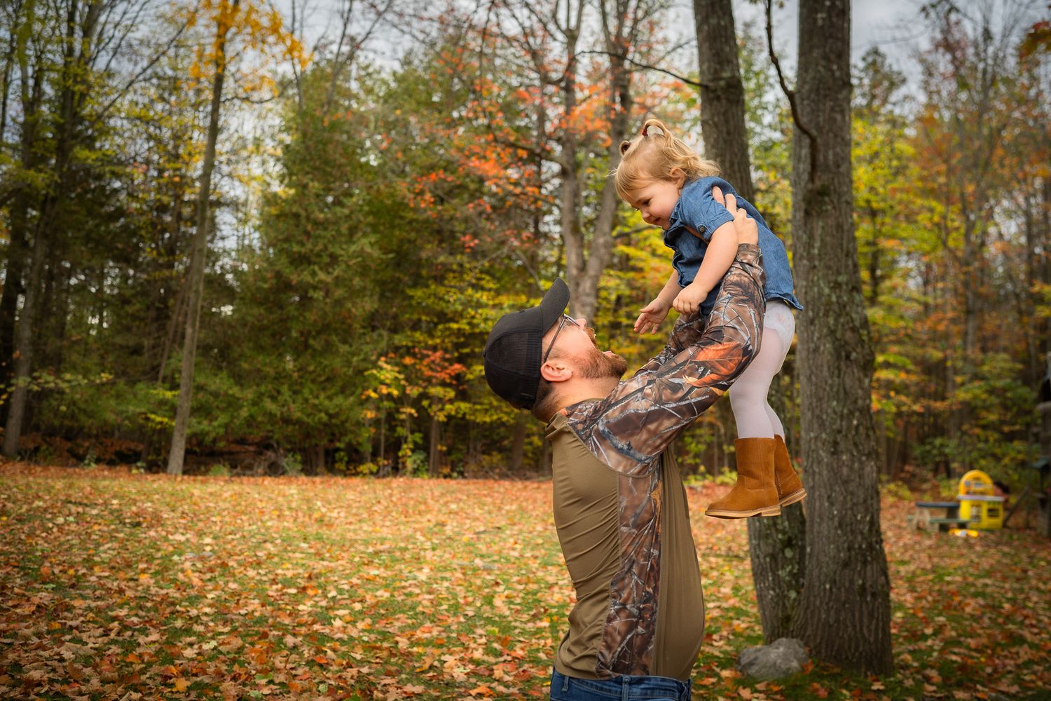 Father lifting toddler in autumn forest park with colorful fall foliage and trees in background.