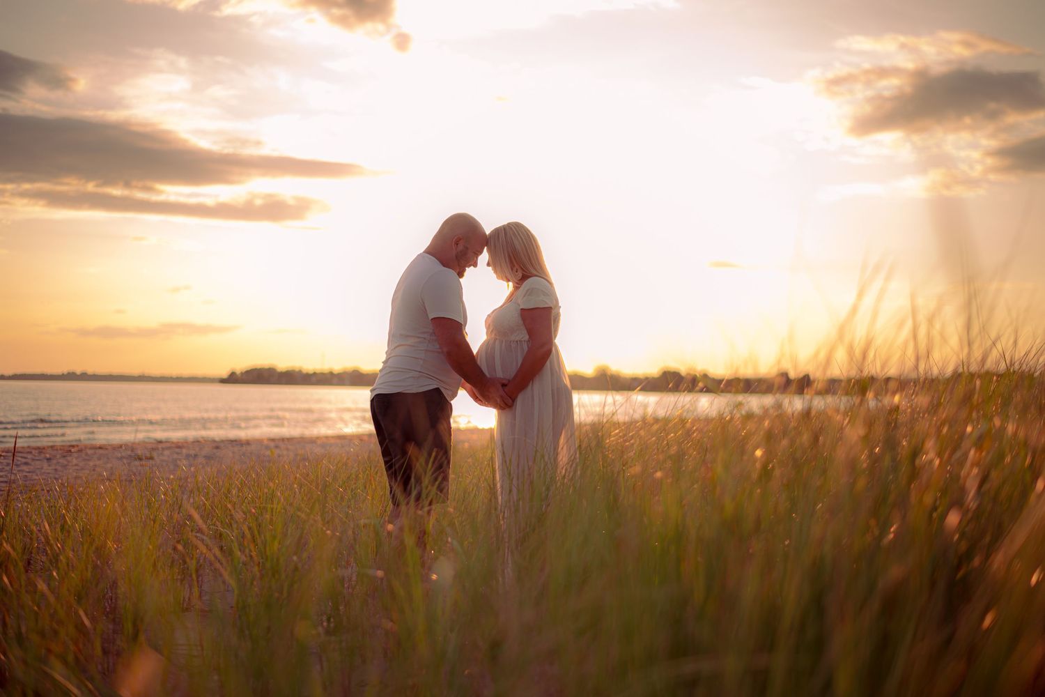 A romantic sunset silhouette of a couple embracing on a beach with tall grass in the foreground. Maternity photographer Ontario