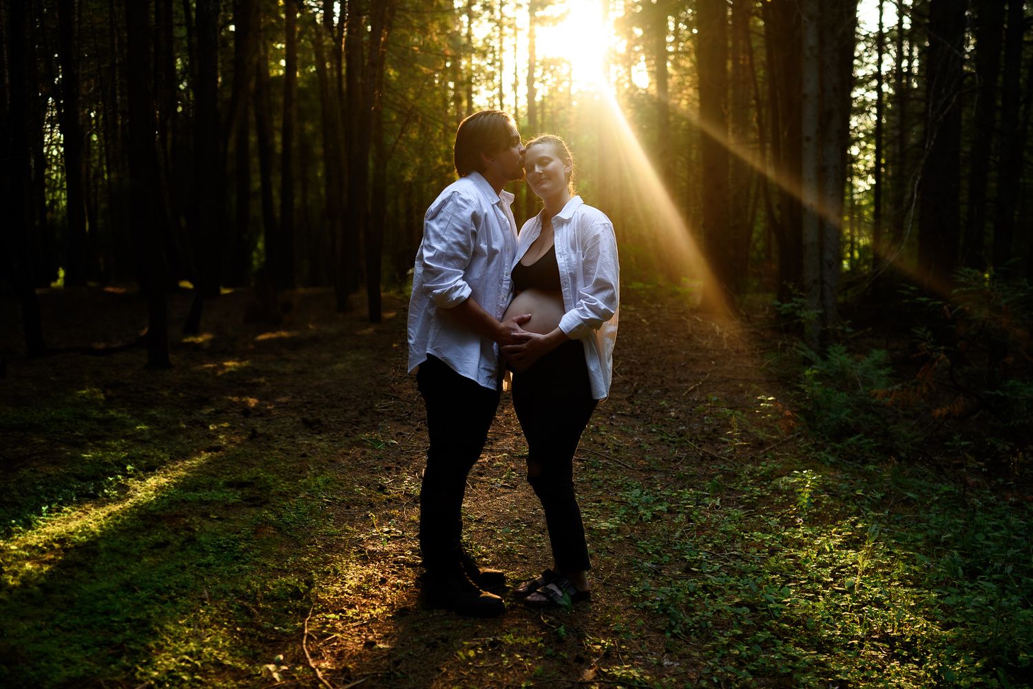 Sunlight streams through a forest creating dramatic beams between the trees during a romantic maternity photoshoot. Maternity photographer O