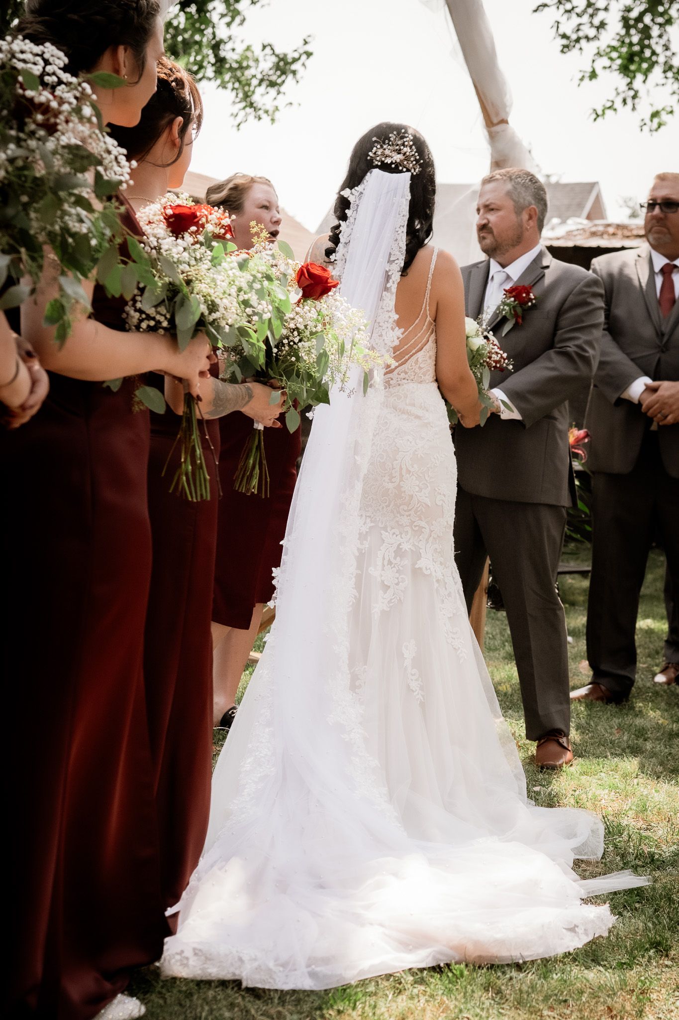 Wedding party stands outdoors with bridesmaids in burgundy dresses and groomsmen in gray suits.