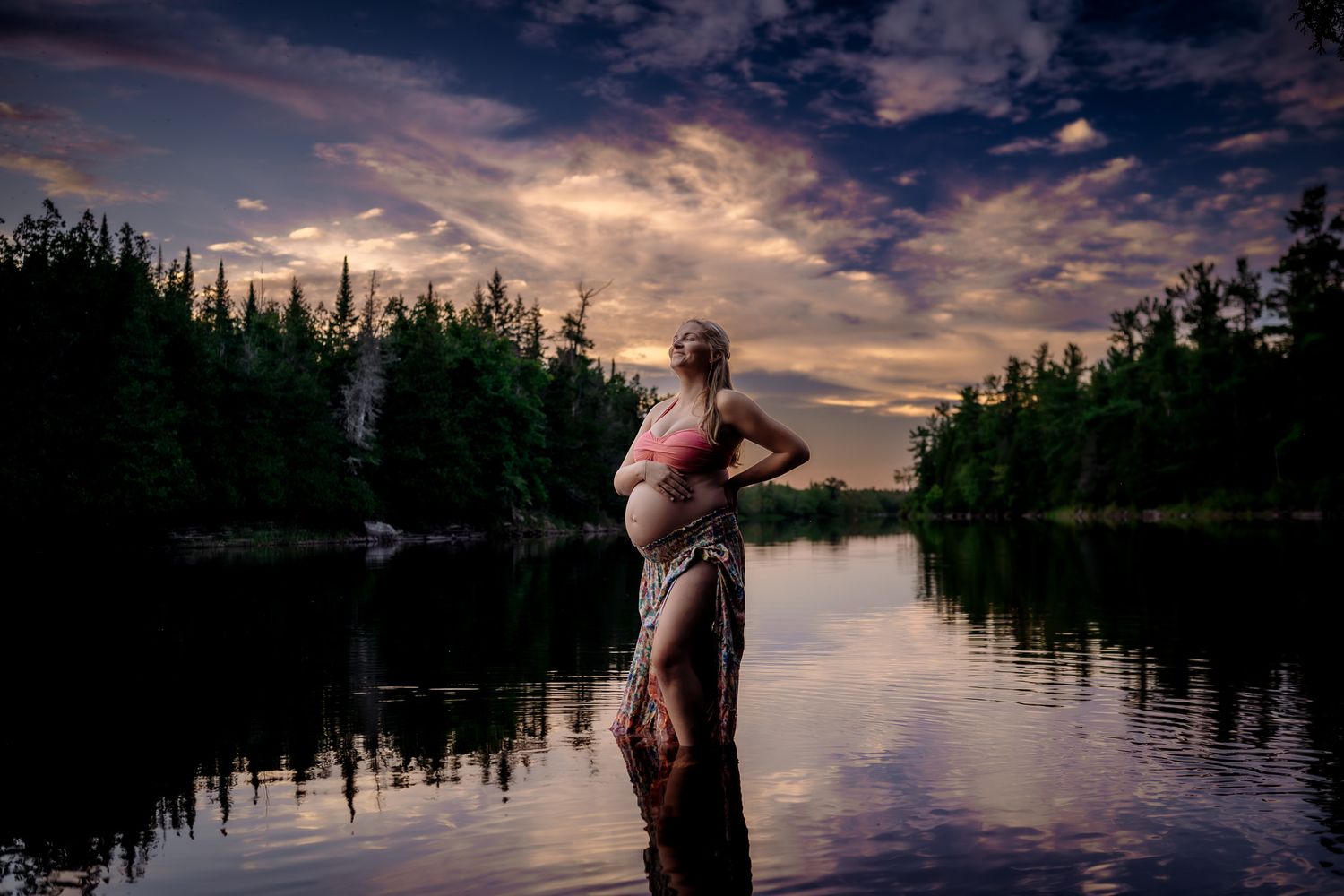 Pregnant woman standing in a serene lake at sunset, surrounded by lush forest and dramatic cloudy sky.