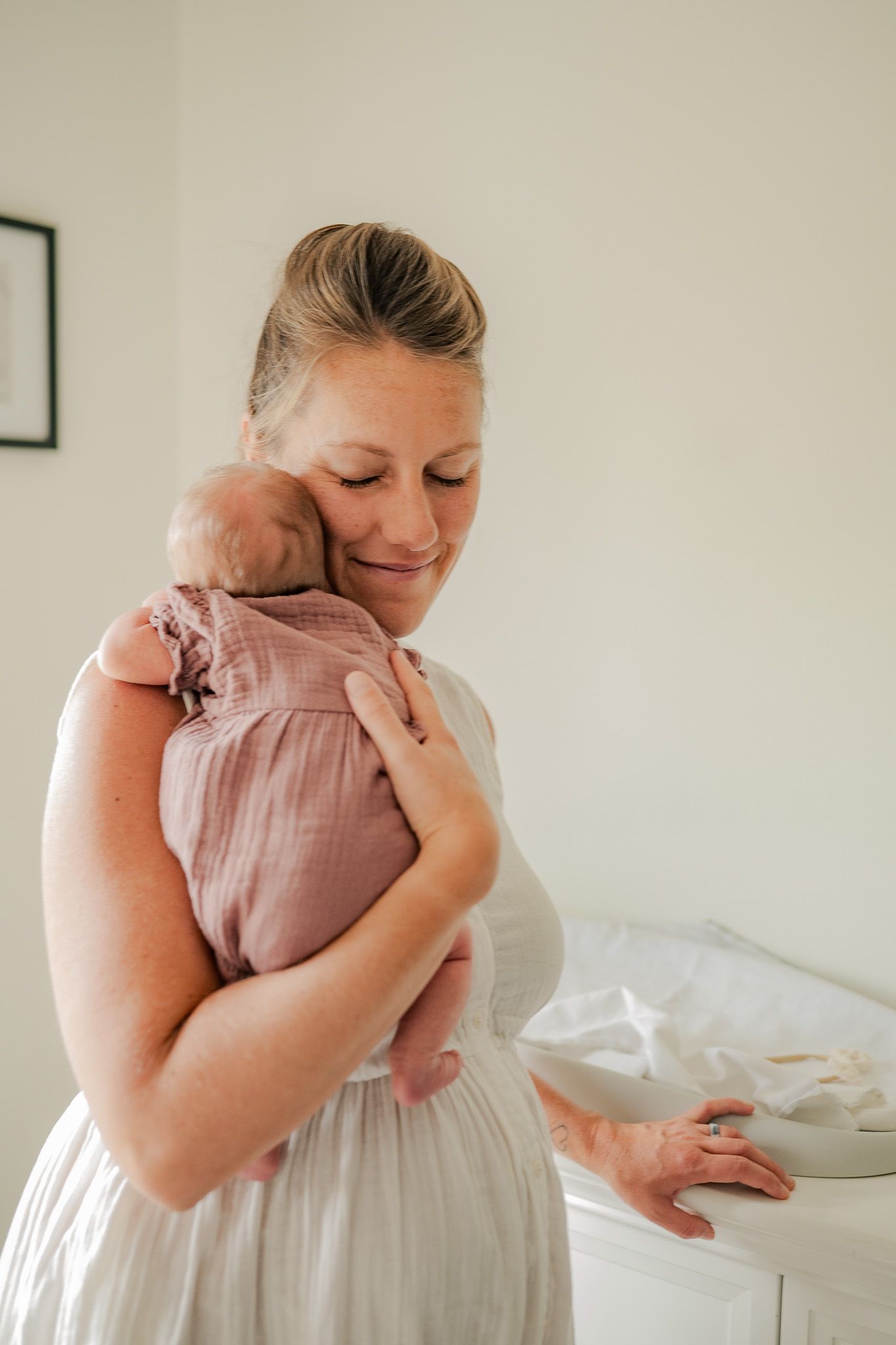 A person in a soft pink dress holds and embraces a small baby in a tender moment against a light background.