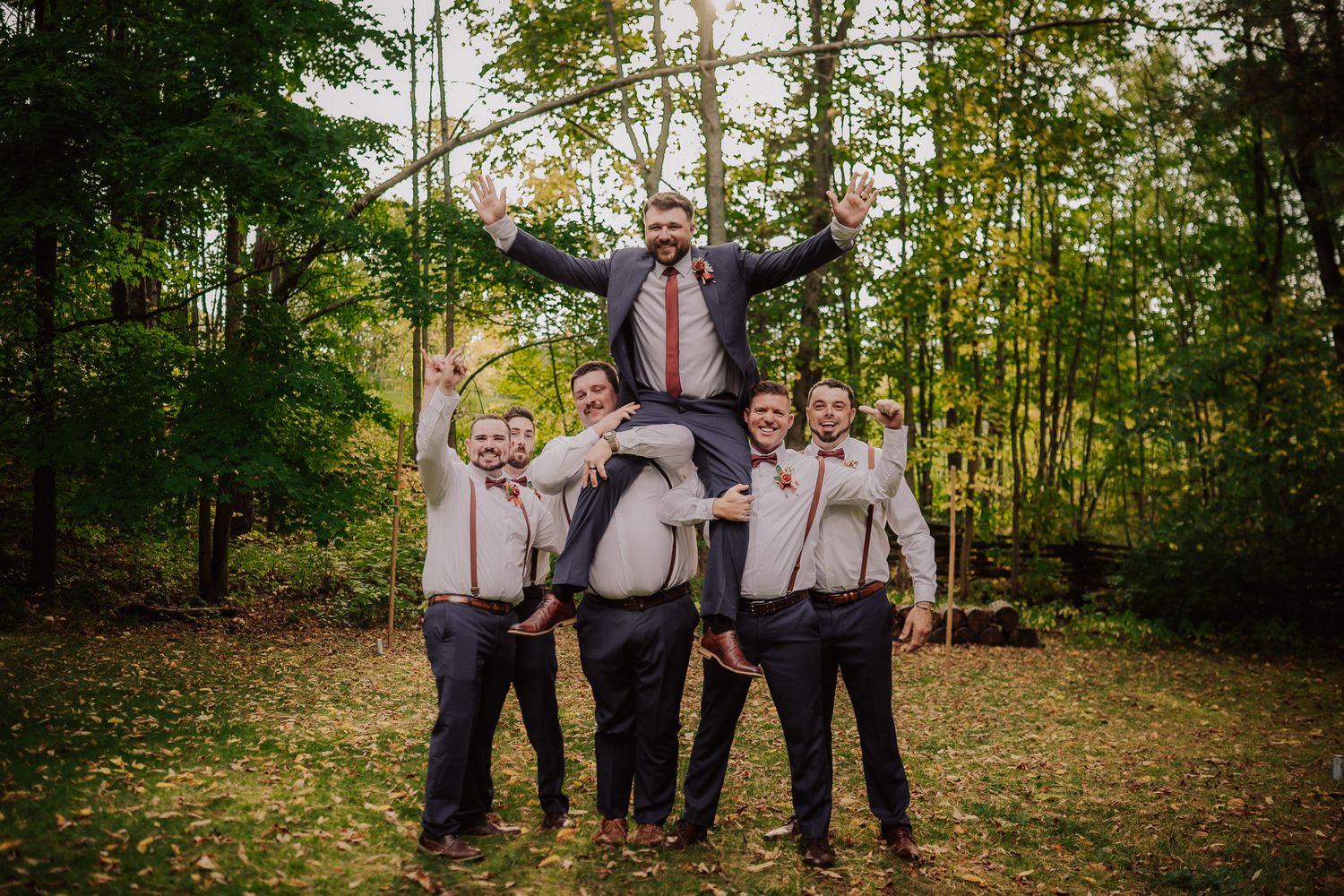 A wedding party in formal attire poses together in a wooded outdoor setting during autumn.