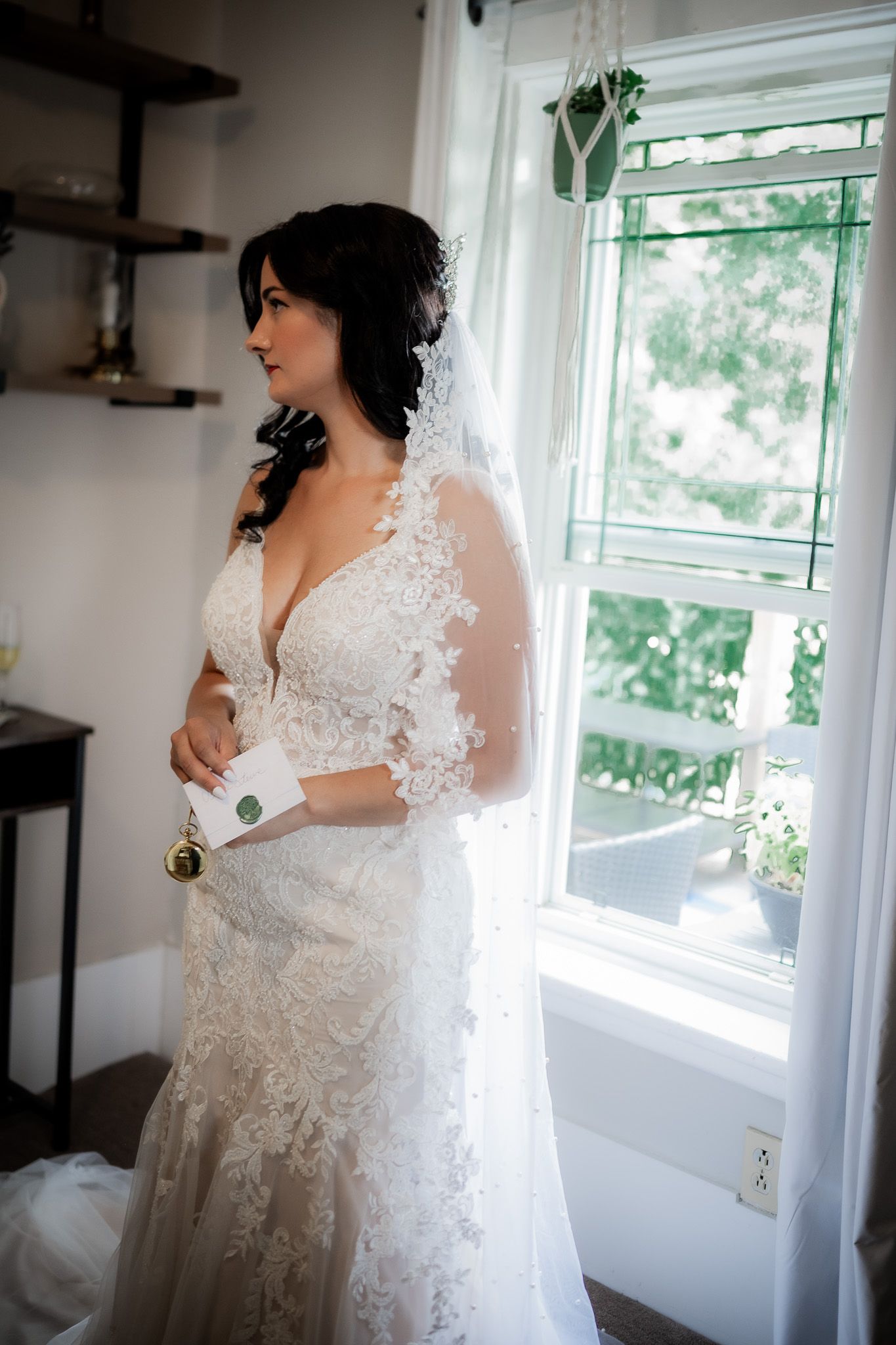 A bride in an elegant white lace wedding dress with long sleeves poses near a bright window in a room.