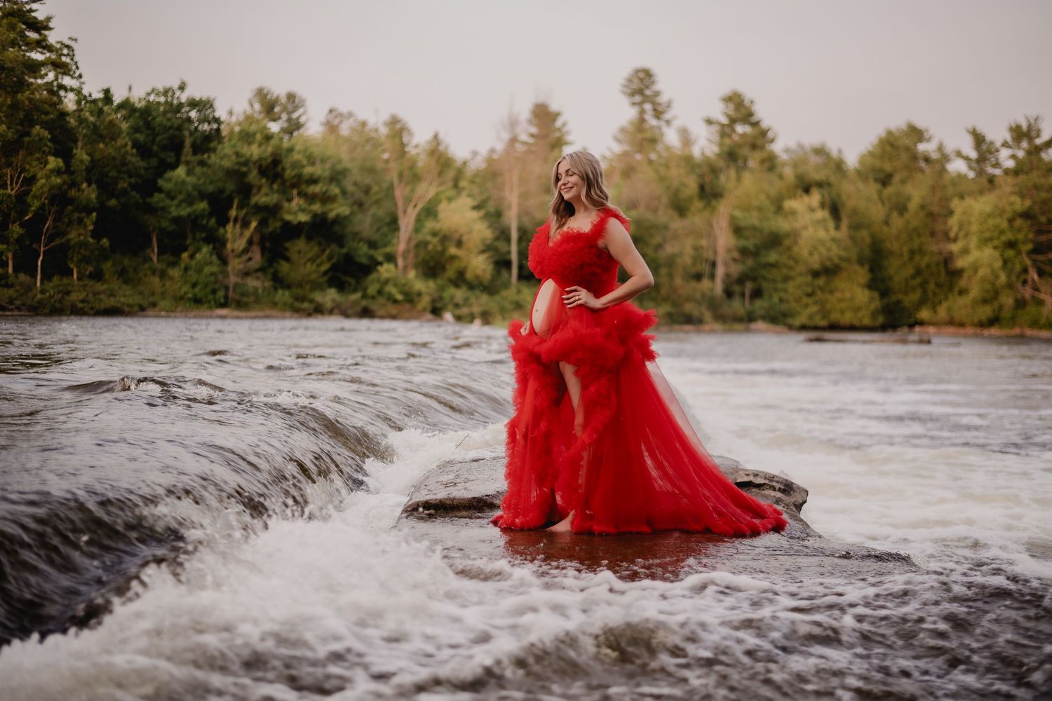 A flowing red dress creates drama against rushing water at the edge of a waterfall. Maternity photographer Ontario