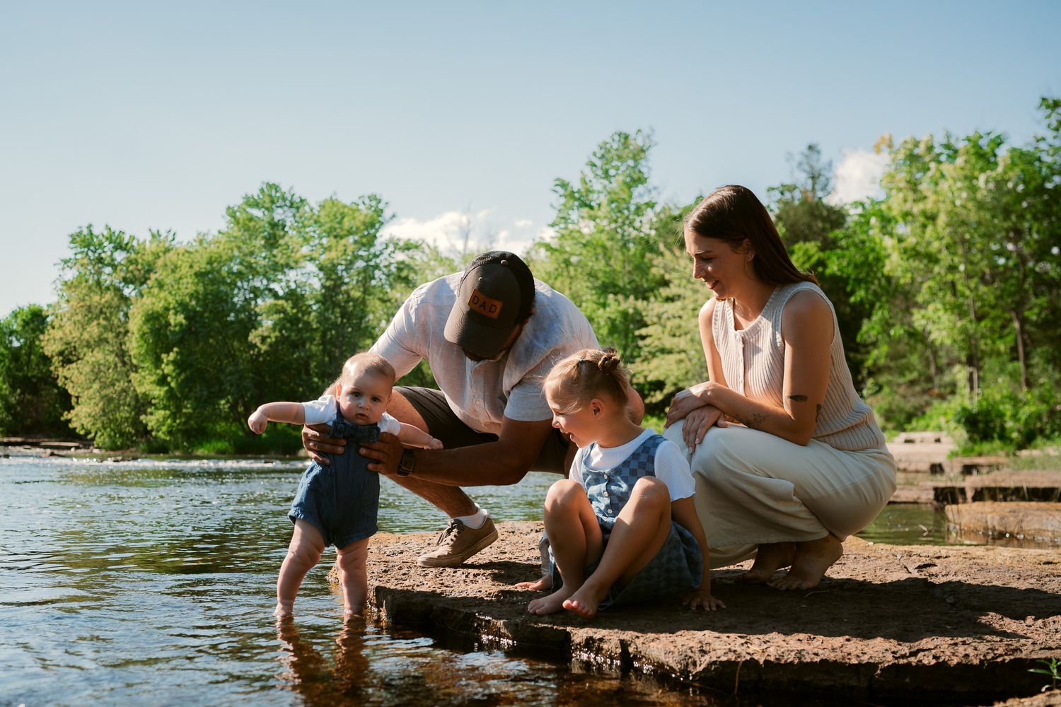 Family enjoying a sunny day by the lake, with parents helping toddler touch the water on a rocky shore.