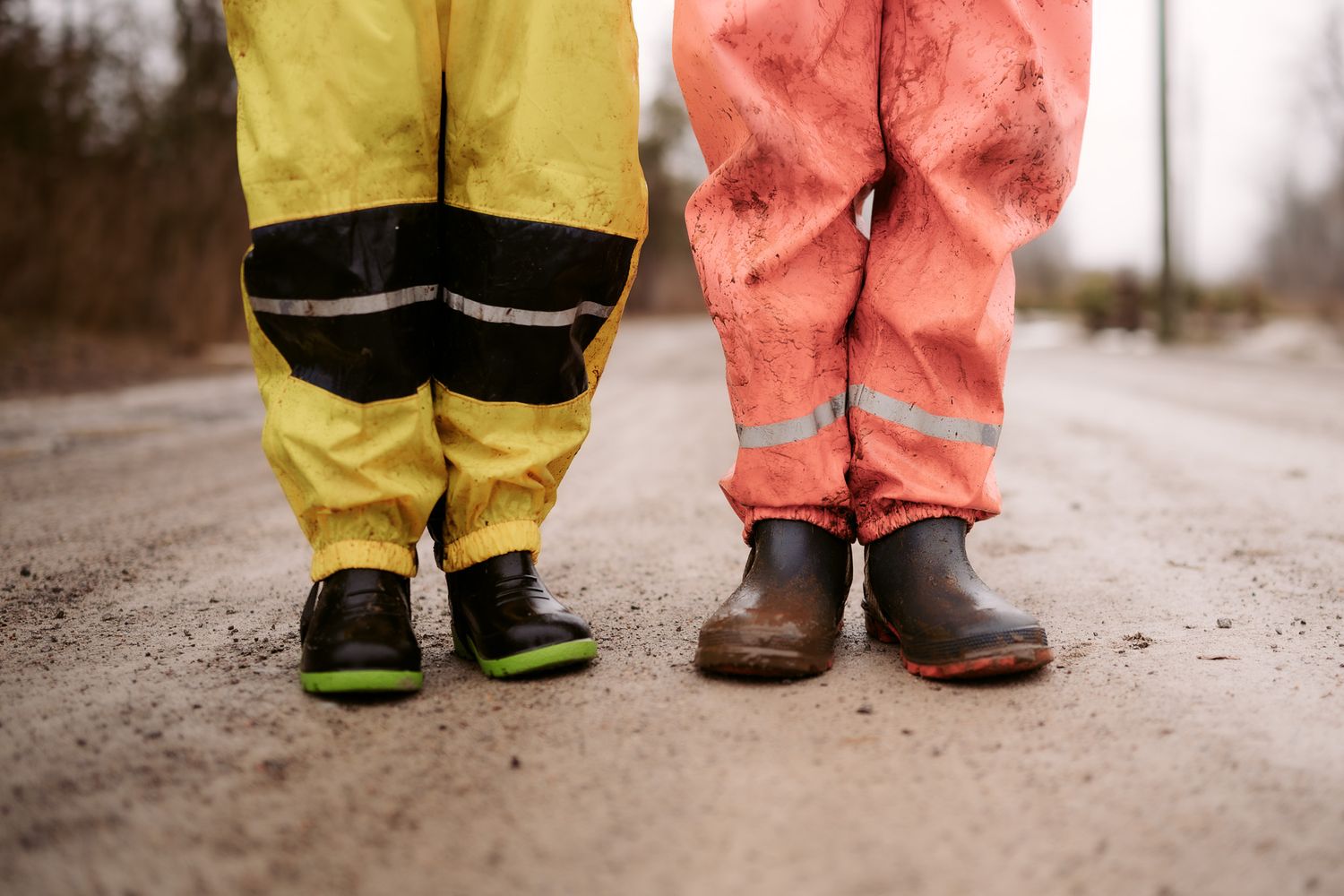 Two children in colorful rain gear and rubber boots standing on a muddy road.