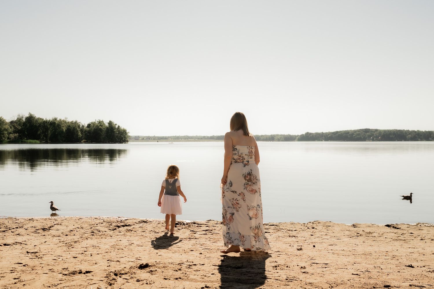 Two people sit peacefully on a sandy beach overlooking a serene lake with trees in the background.