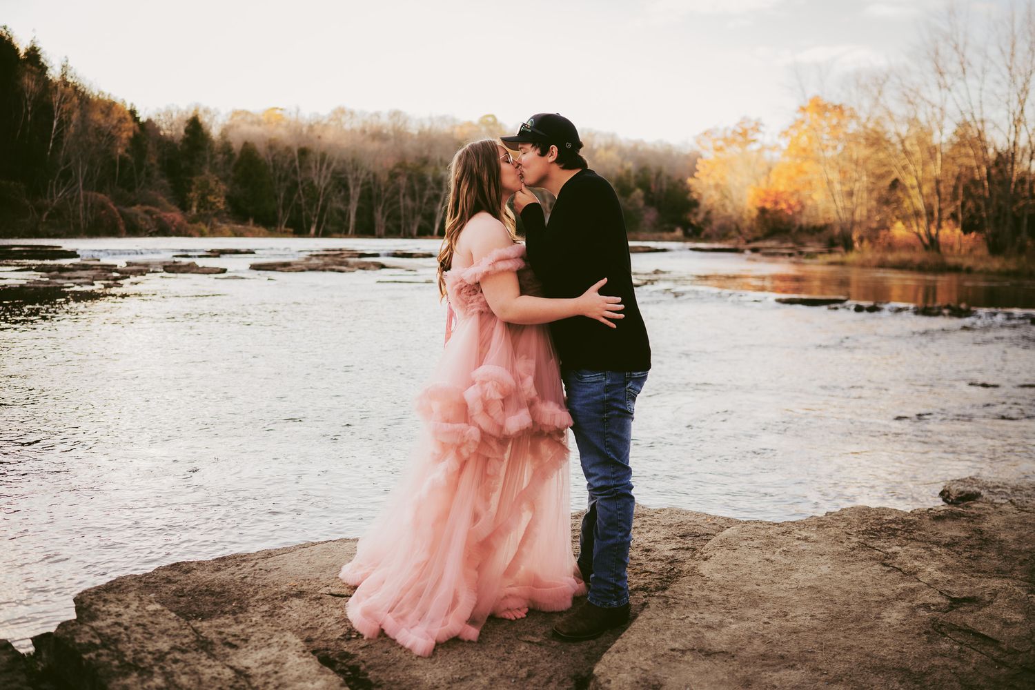 Couple kissing by a river in autumn, woman in pink ruffled gown, man in black jacket, standing on rocks at golden hour.