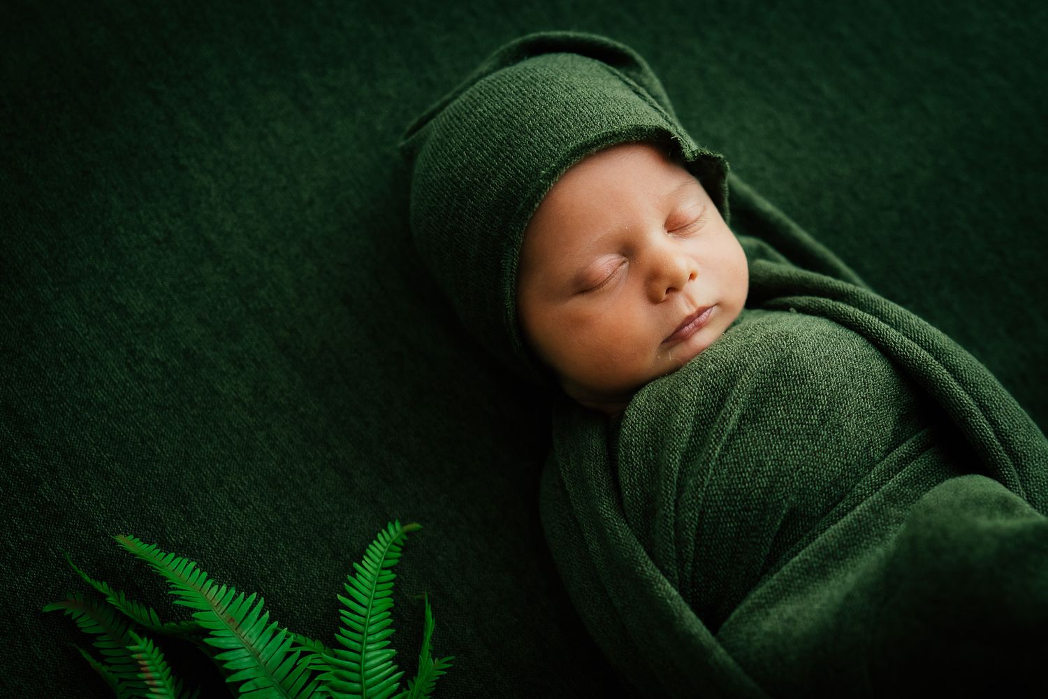 Sleeping newborn wrapped in dark green fabric against matching background.