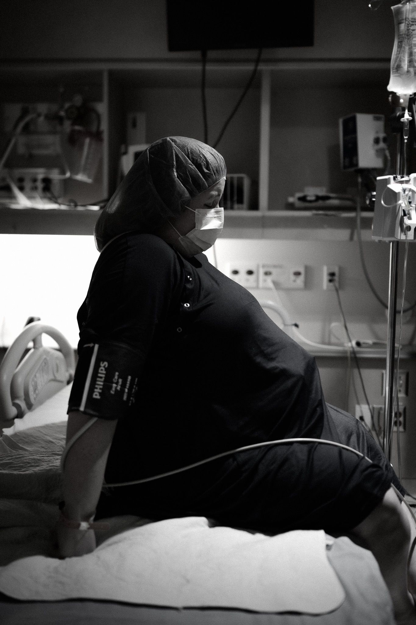 Pregnant woman in black scrubs and mask sits on hospital bed with blood pressure cuff, IV equipment visible.