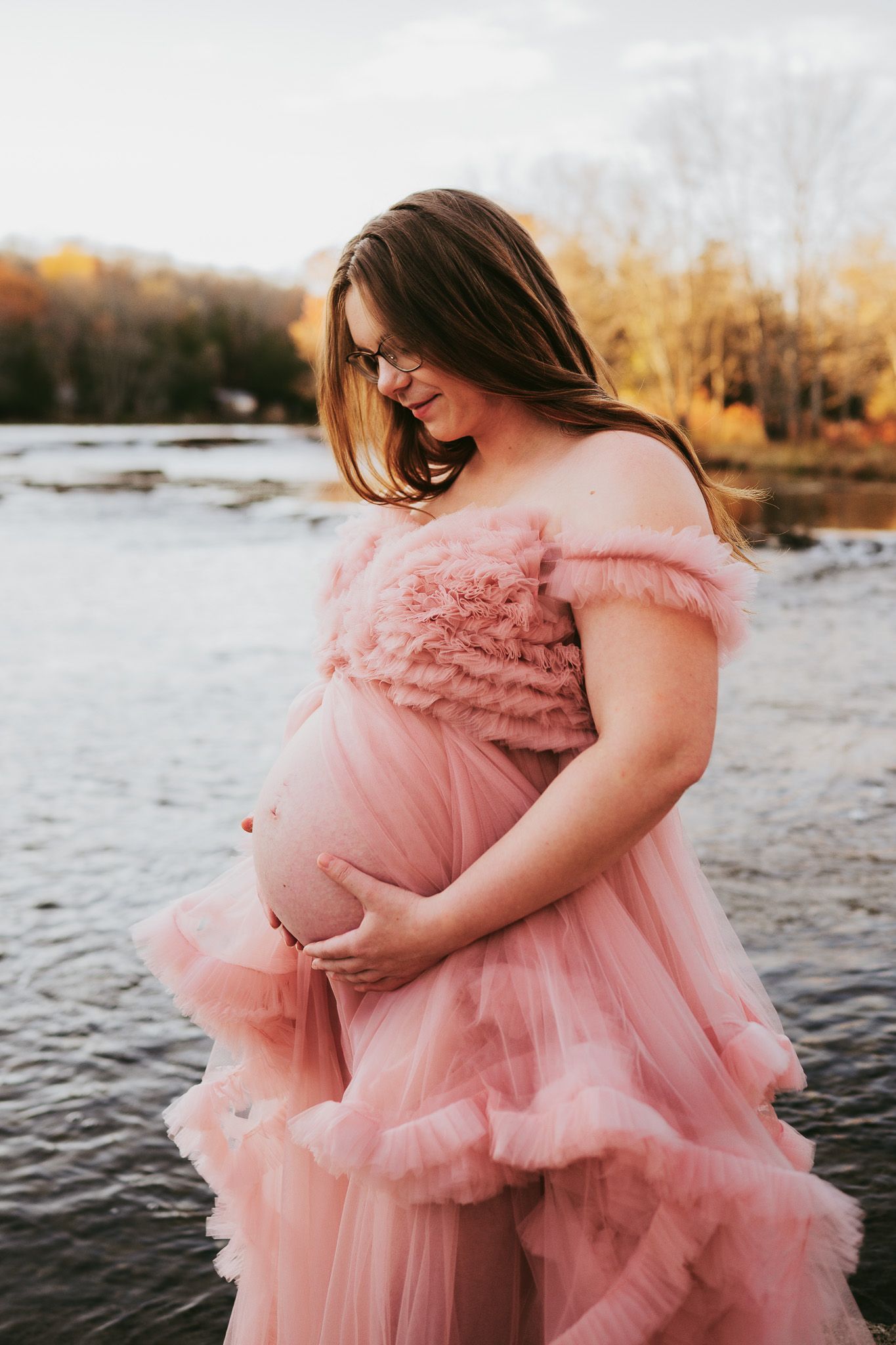 Expectant mother in flowing pink tulle gown poses by a lake during golden hour sunset. Maternity photographer Ontario
