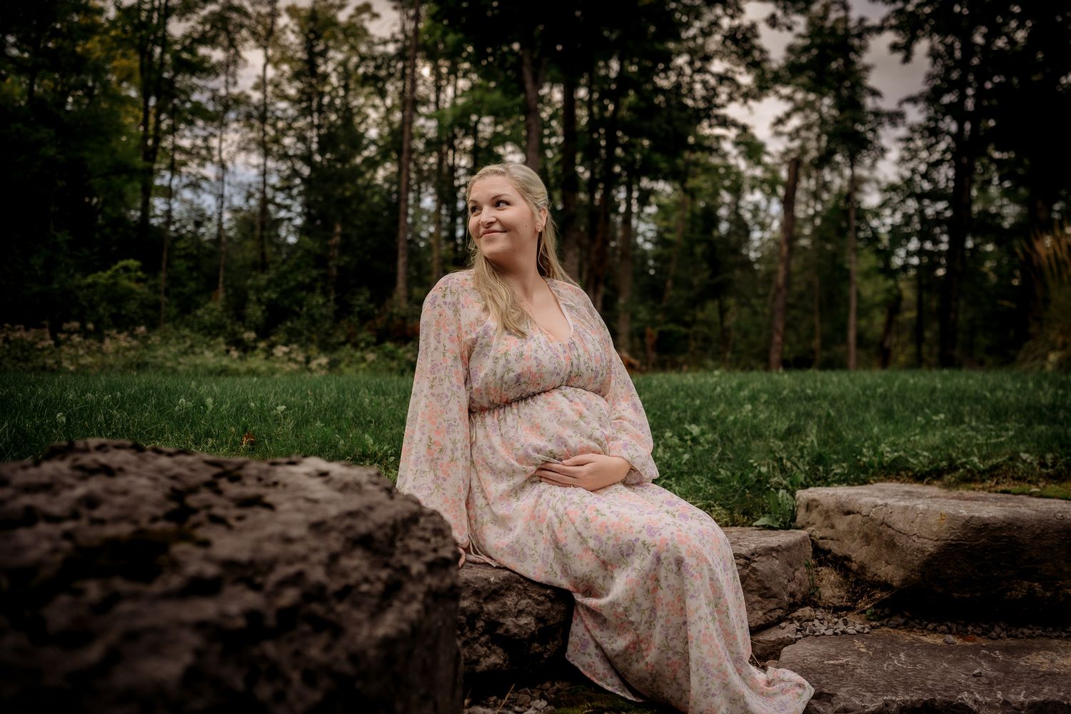 Pregnant woman in floral dress sitting on rocks in a lush green forest during a maternity photo session.