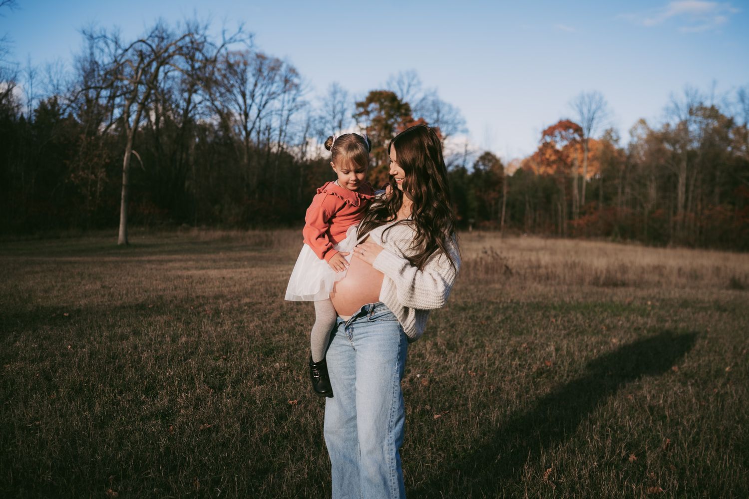 Someone in light jeans holds a child while standing in a grassy field at sunset with bare trees in the background.