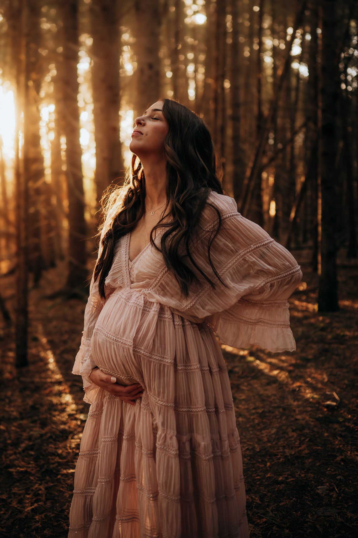 A person in a flowing pink dress poses ethereally in a sunlit forest during golden hour. Maternity photographer Ontario