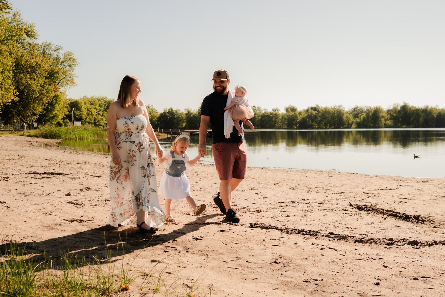 A family walks together on a sandy beach at sunset beside a tranquil lake with trees in the background.