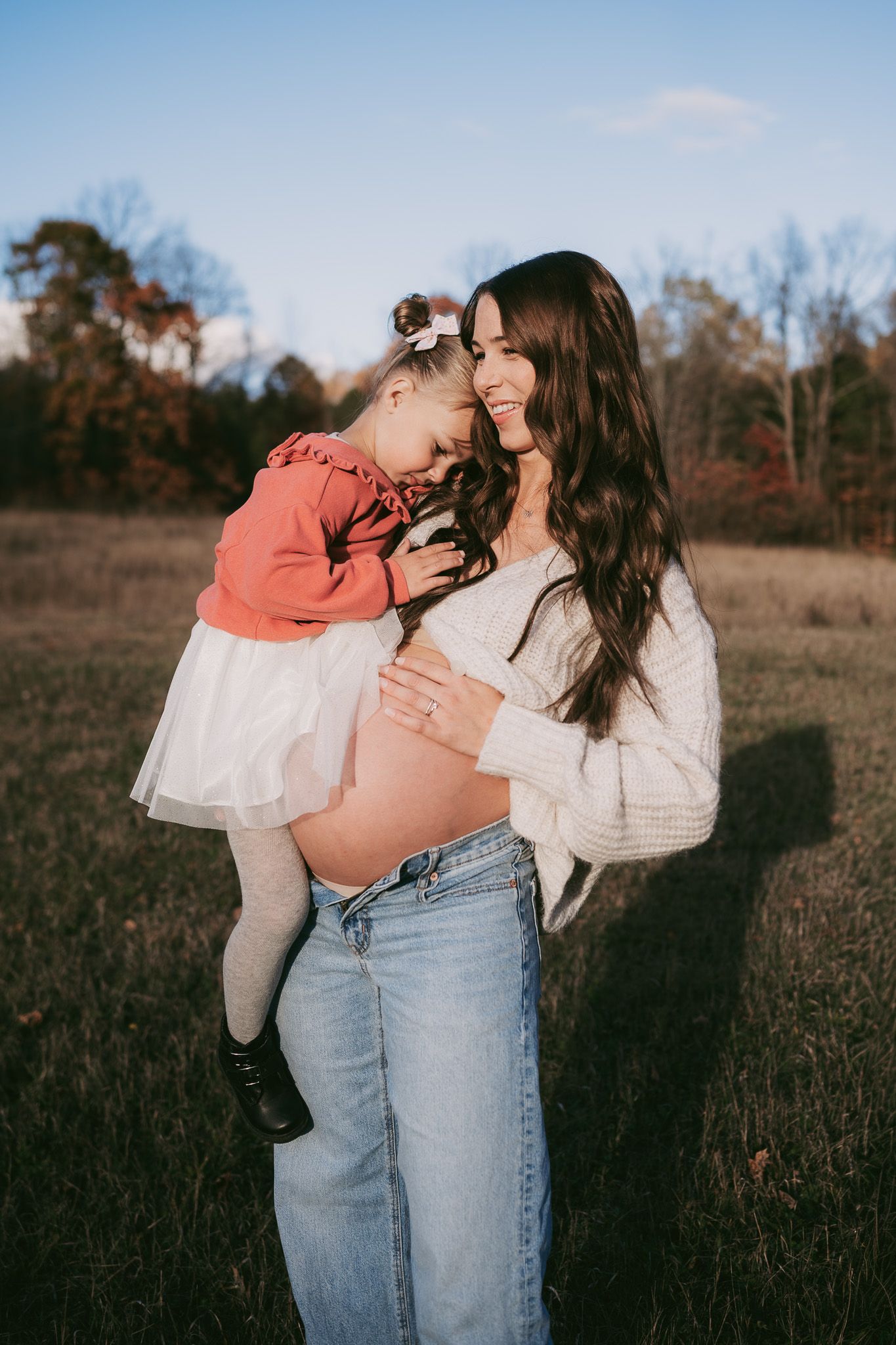 A couple shares an intimate moment in a field at sunset wearing light colored clothing and denim jeans. Maternity photographer Ontario