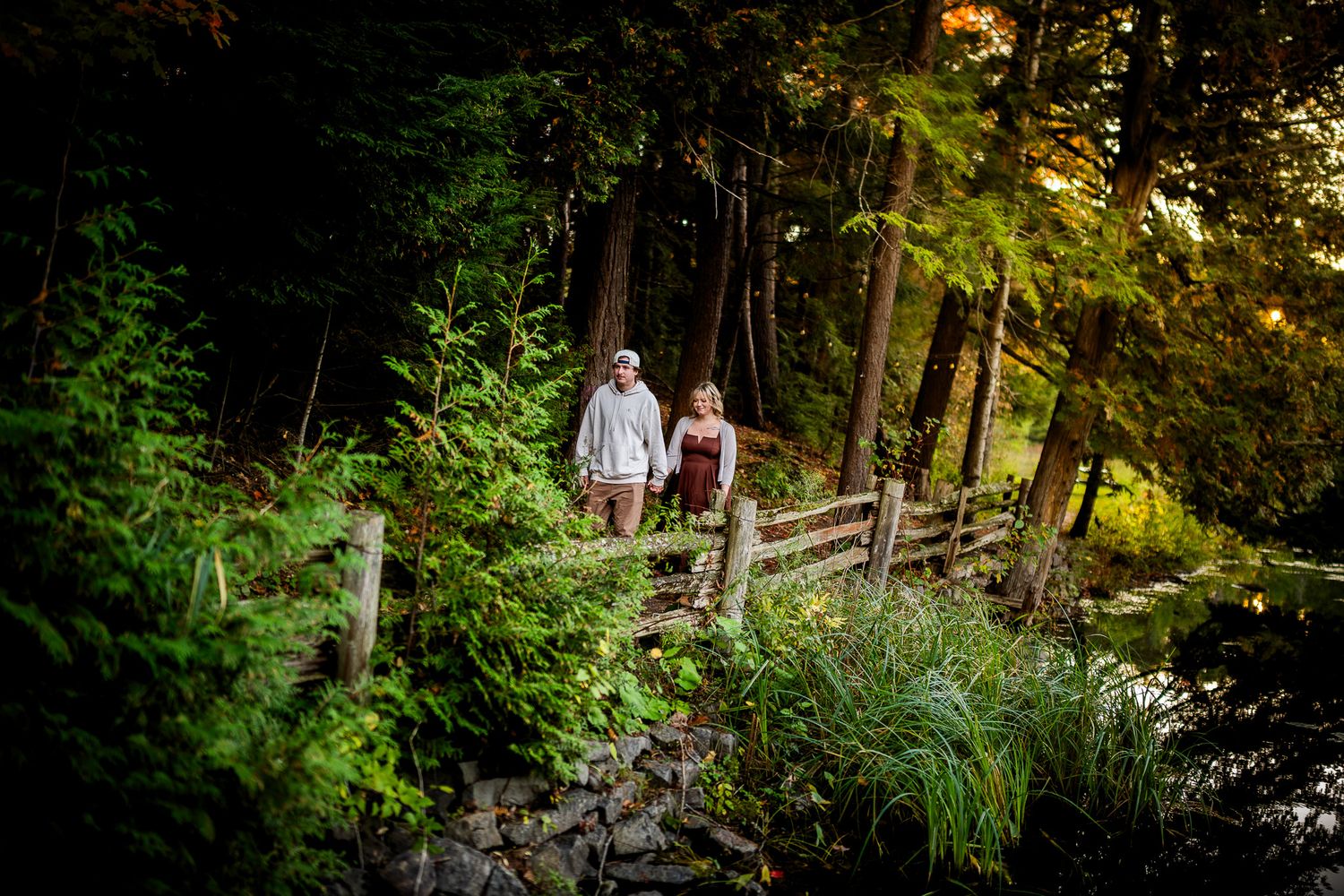 Couple holding hands walking on a wooden bridge surrounded by lush green forest trees in golden hour light.