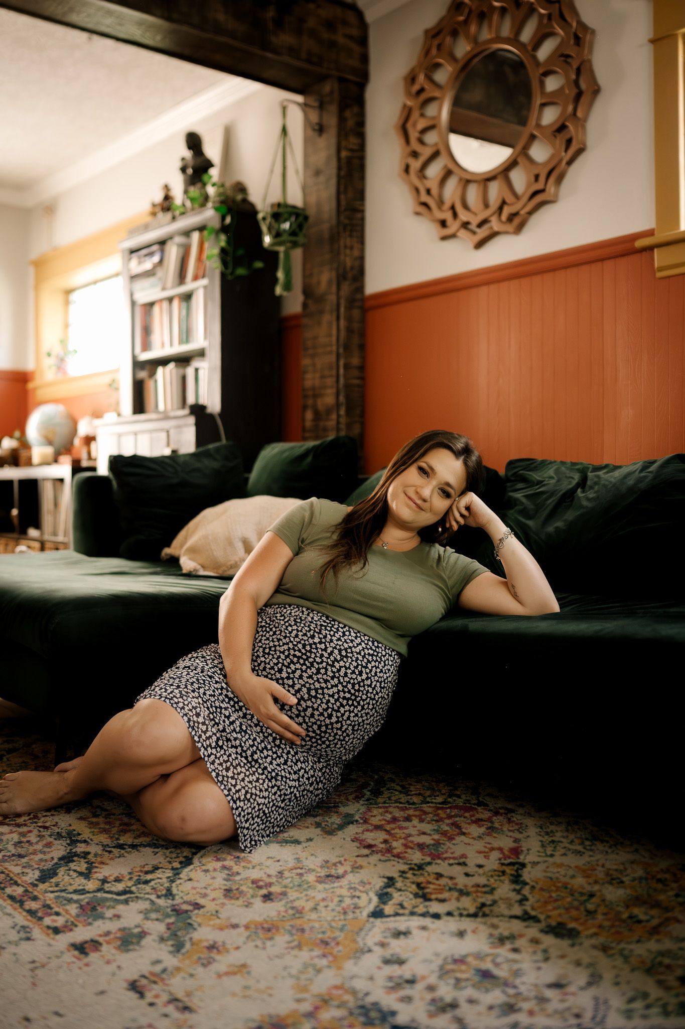 Pregnant woman in olive top and leopard skirt relaxing on floor against green velvet sofa in cozy living room.
