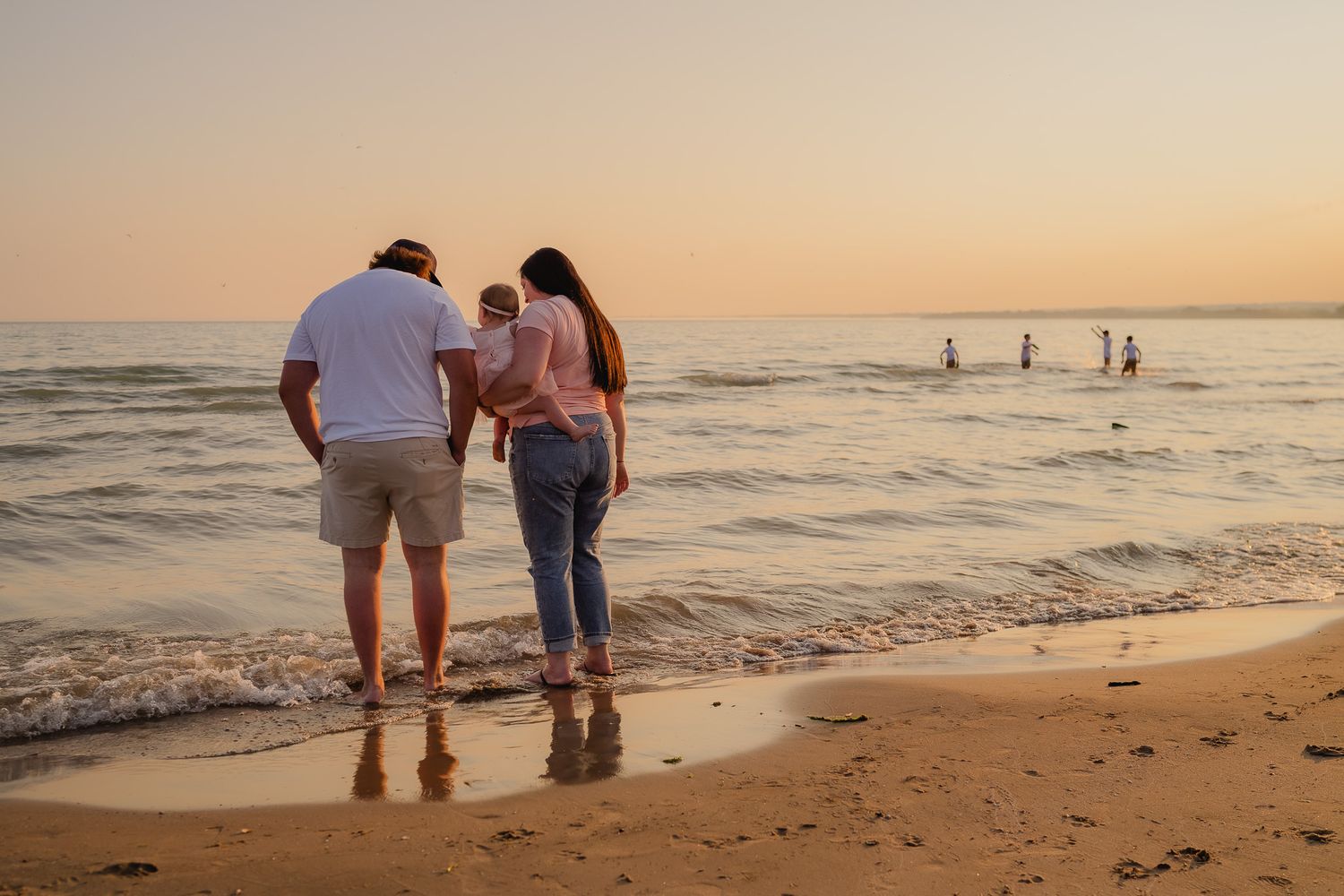 Family of three standing at the shoreline during a golden sunset on a sandy beach.