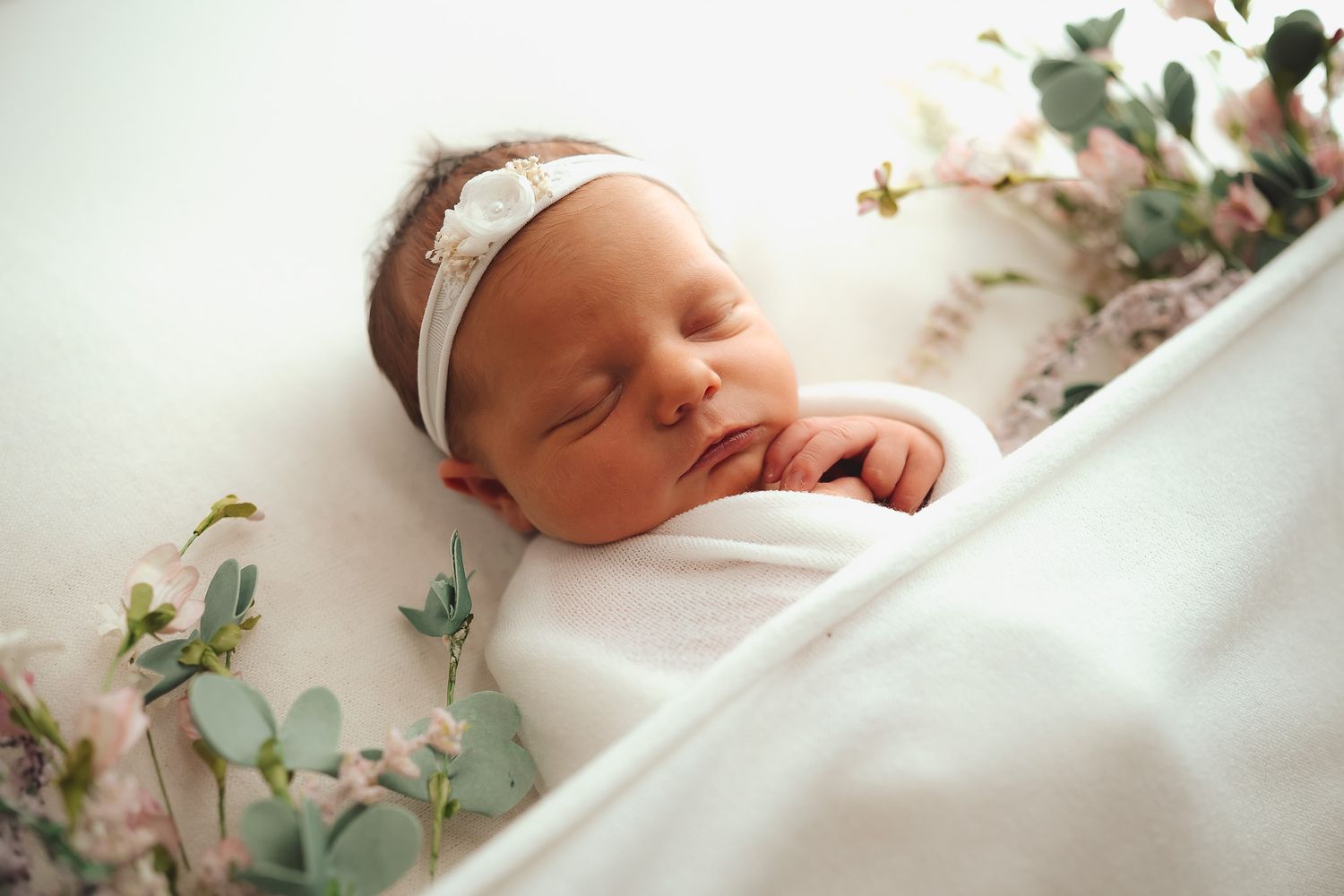 Delicate floral arrangements frame a peaceful sleeping newborn wrapped in soft white fabric.