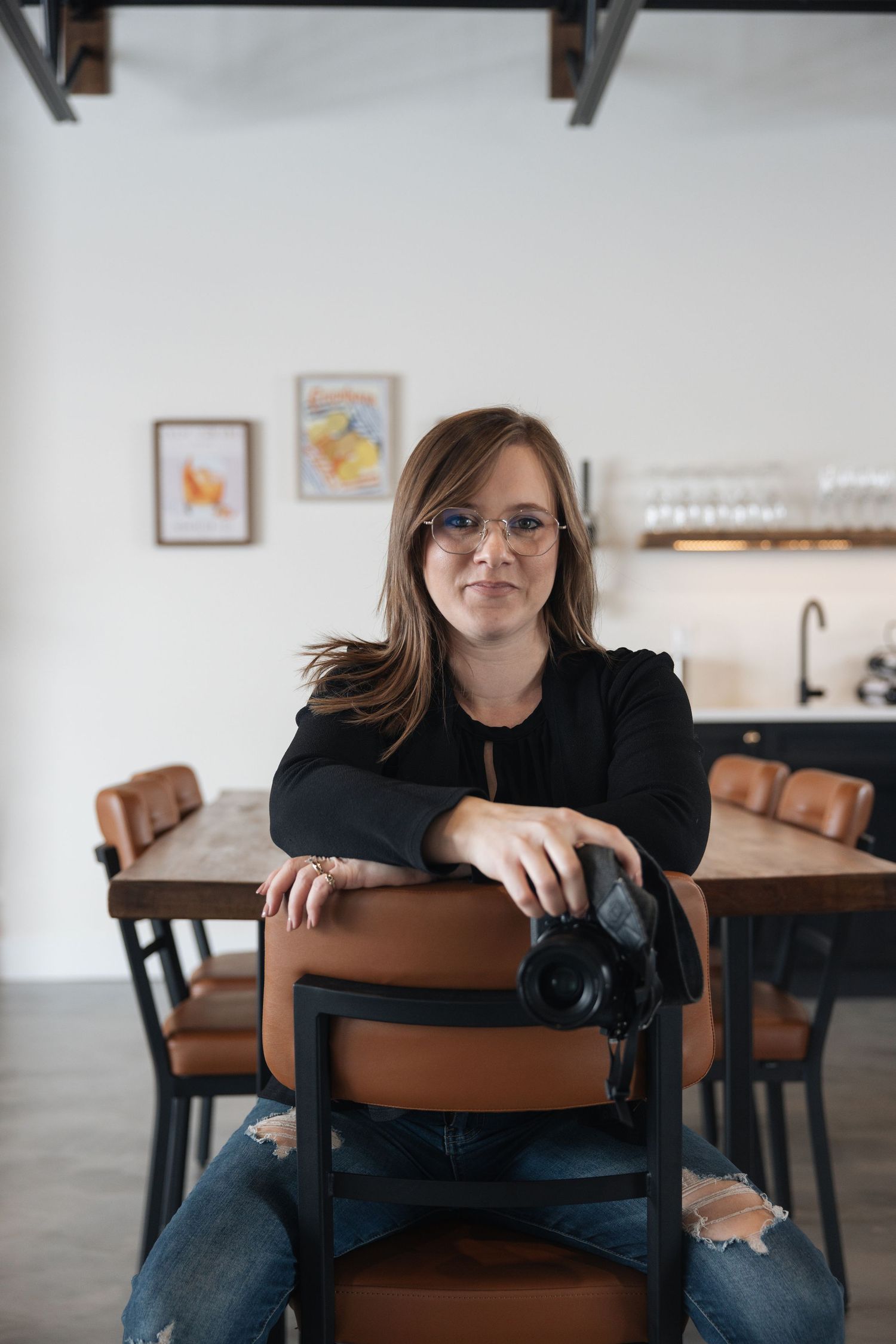 A person in black clothing leans on a wooden chair in a minimalist room with white walls and framed artwork.