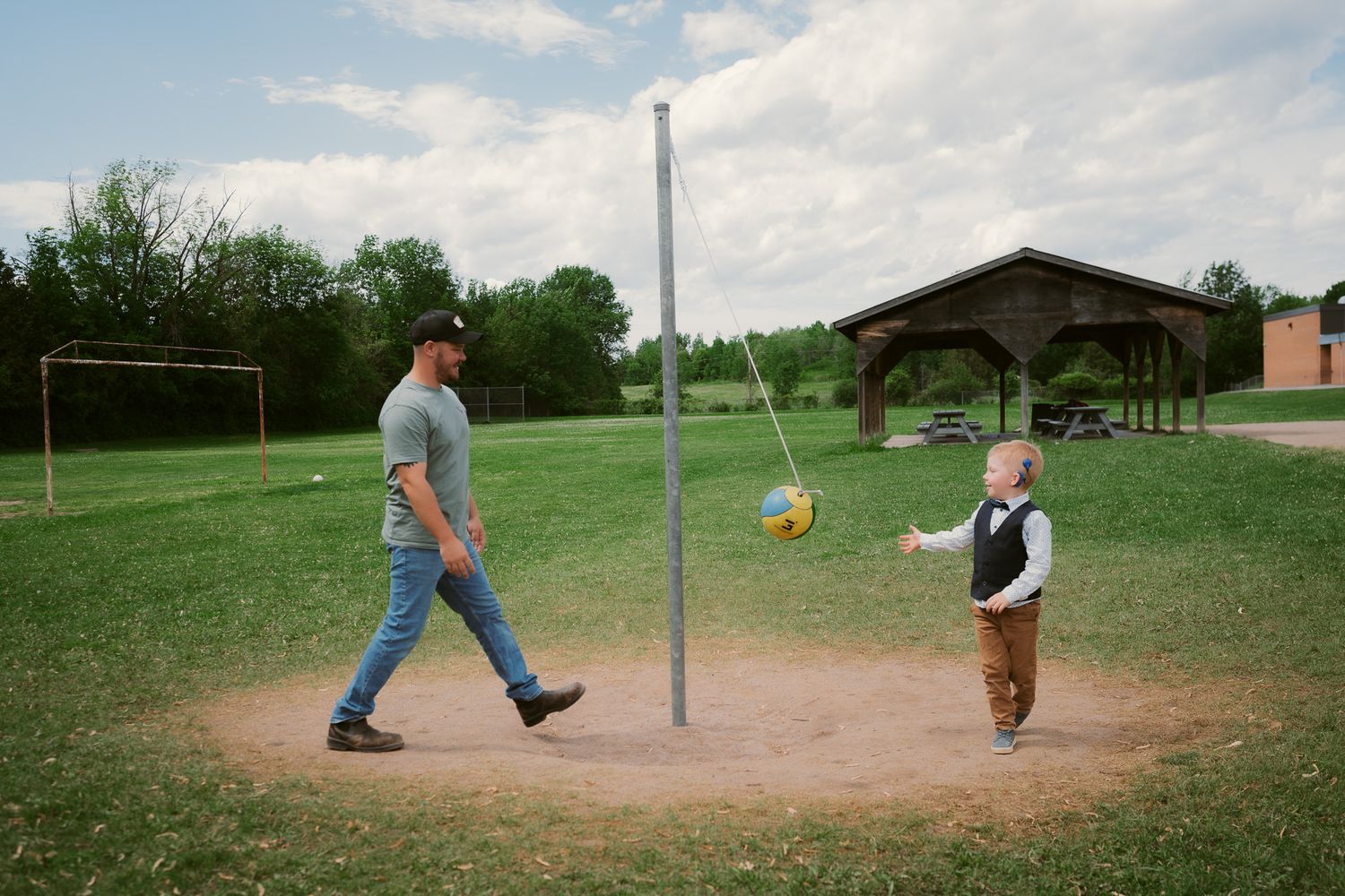 Man and young boy playing tetherball on a grassy field near a wooden gazebo on a cloudy day.