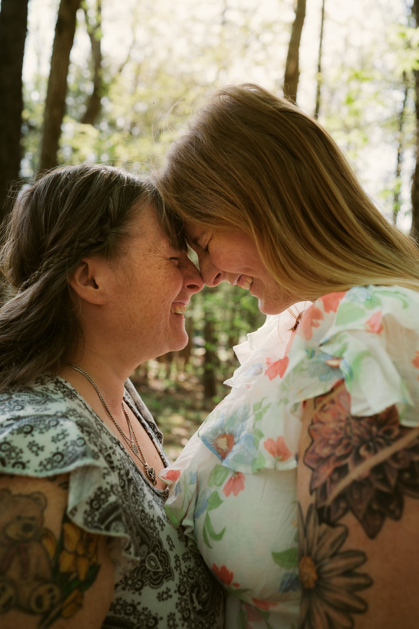 Two women with tattoos touch foreheads and smile in a sun-dappled forest, sharing an intimate moment.
