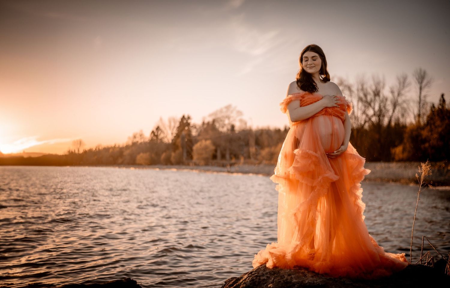A flowing peach-colored gown creates an ethereal scene against a sunset waterfront backdrop. Maternity photographer Ontario
