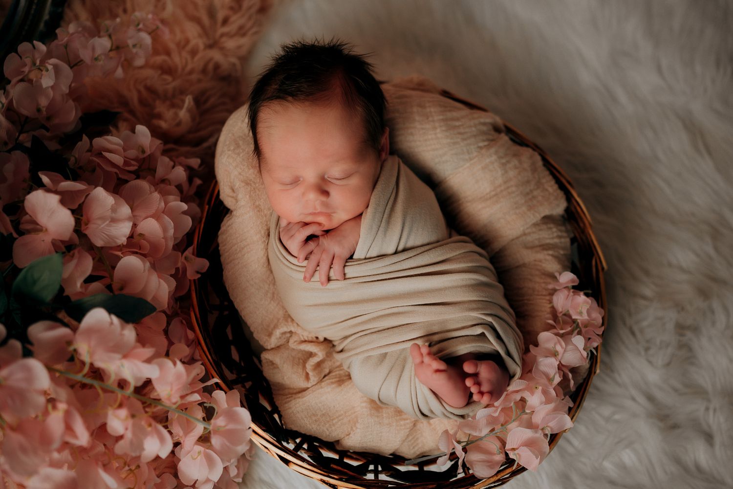 Newborn baby swaddled in cream wrap sleeping in a basket surrounded by pink flowers on a white fur background.