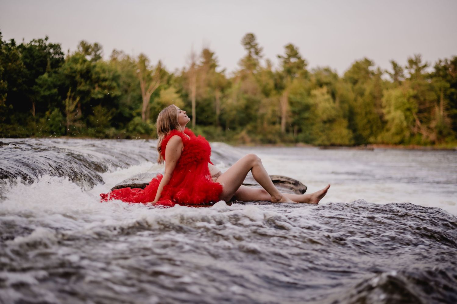 Woman in red dress sitting on rocky waterfall edge with lush green trees in background.