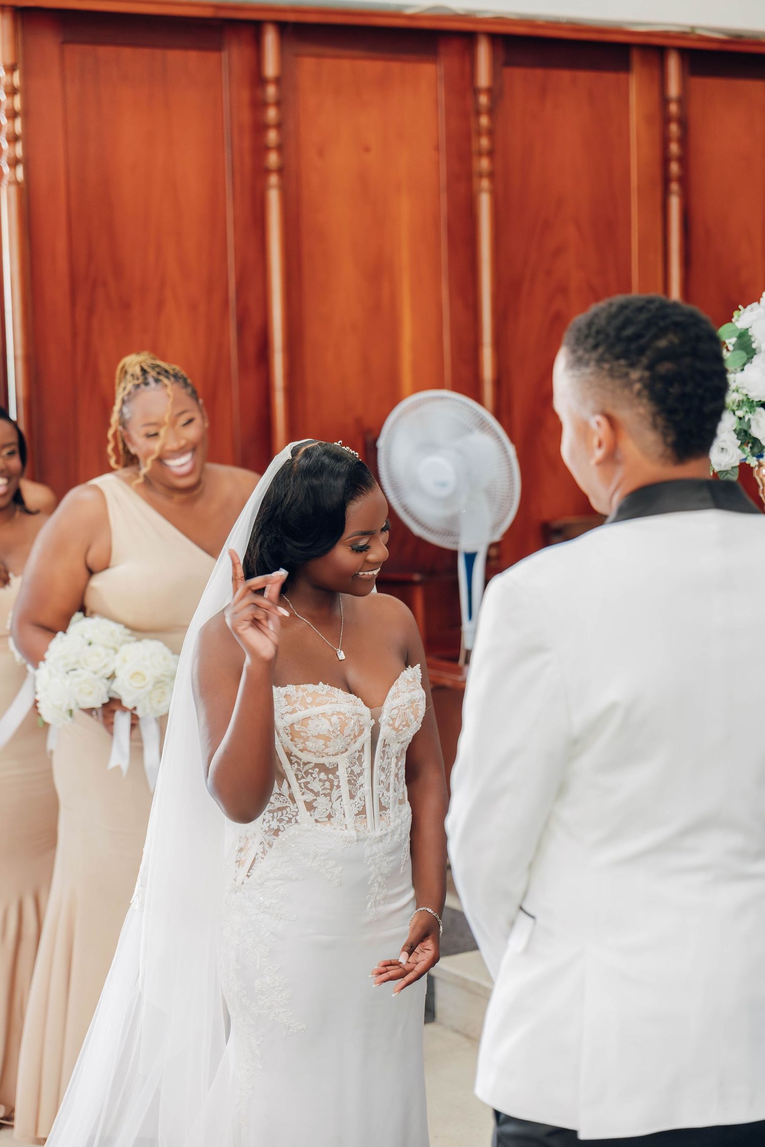Bride in lace wedding gown and veil smiles at groom in white tuxedo during wedding ceremony with bridesmaids behind her.