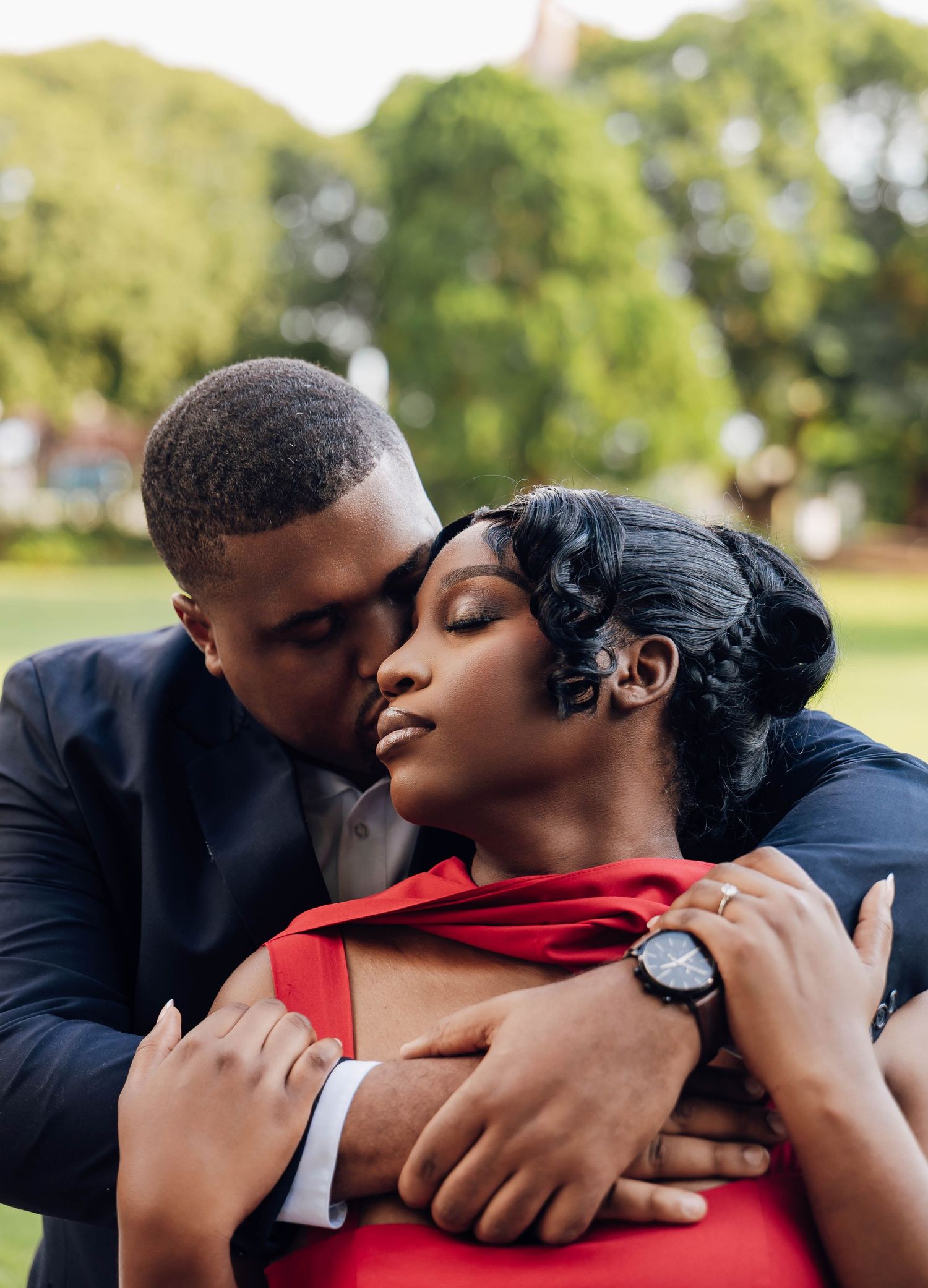 Romantic couple embracing outdoors, man in navy suit kissing woman in red dress on the forehead in a lush green park.
