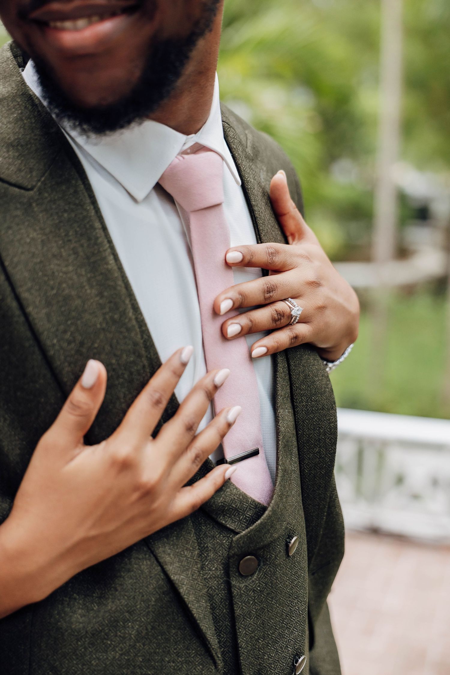 Close-up of bride's hands with diamond ring resting on groom's grey suit with pink tie.
