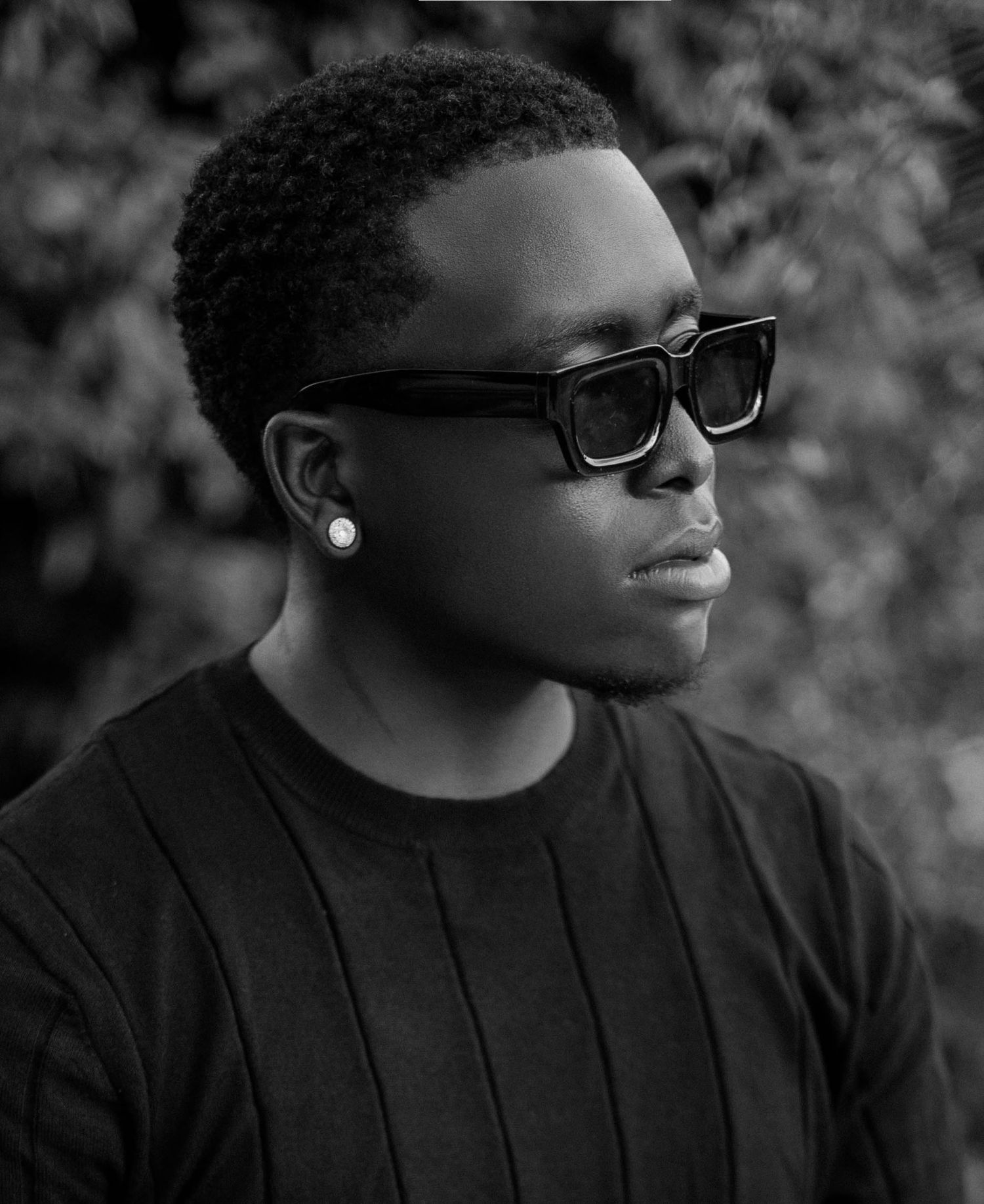 Black and white portrait of a Ah-keel Richard Mitchell wearing sunglasses and a diamond earring, dressed in a ribbed black sweater.