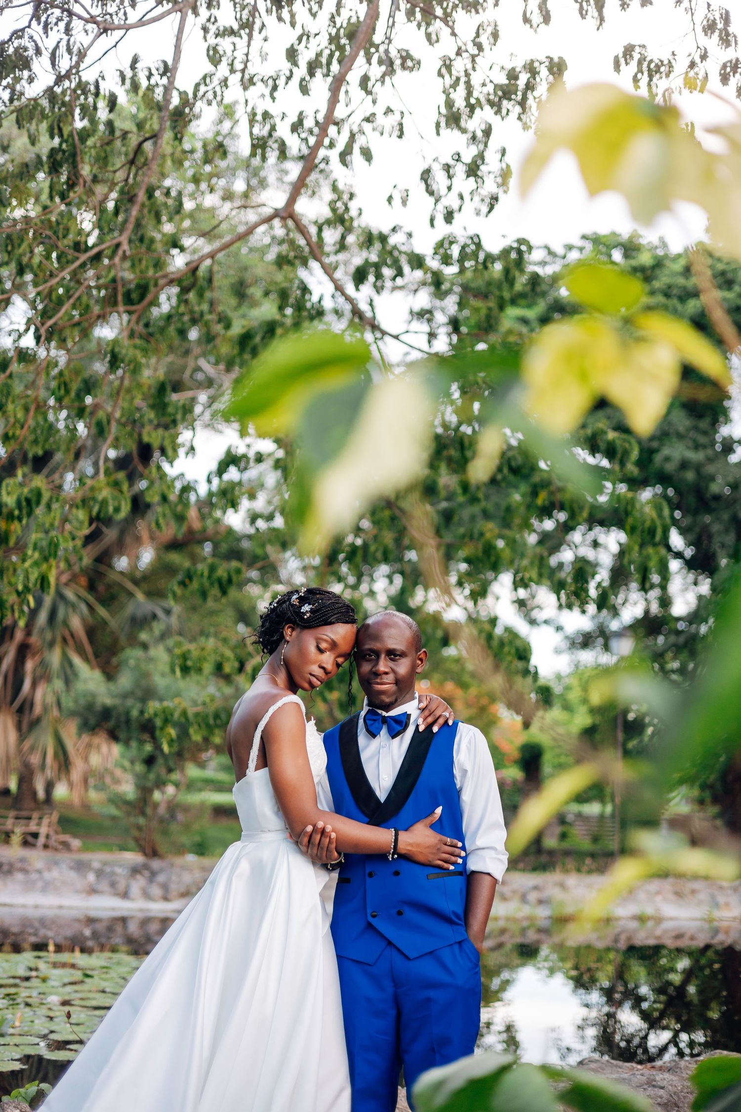 Black bride and groom in blue suit embracing near a pond surrounded by lush greenery, sharing a tender wedding moment.